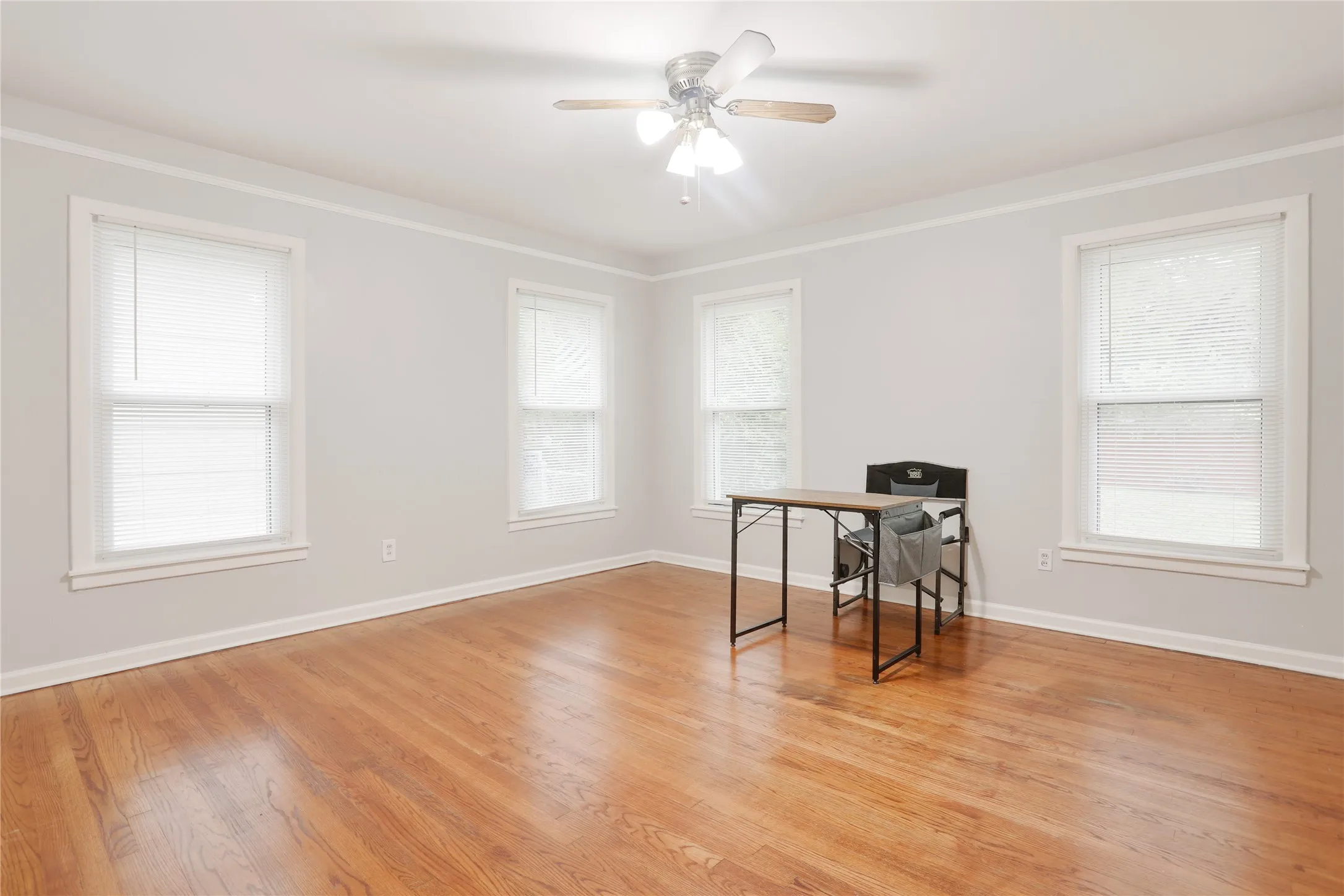 Warm wood-finish flooring, multiple windows with white trim, a ceiling fan with light fixture, and crown molding