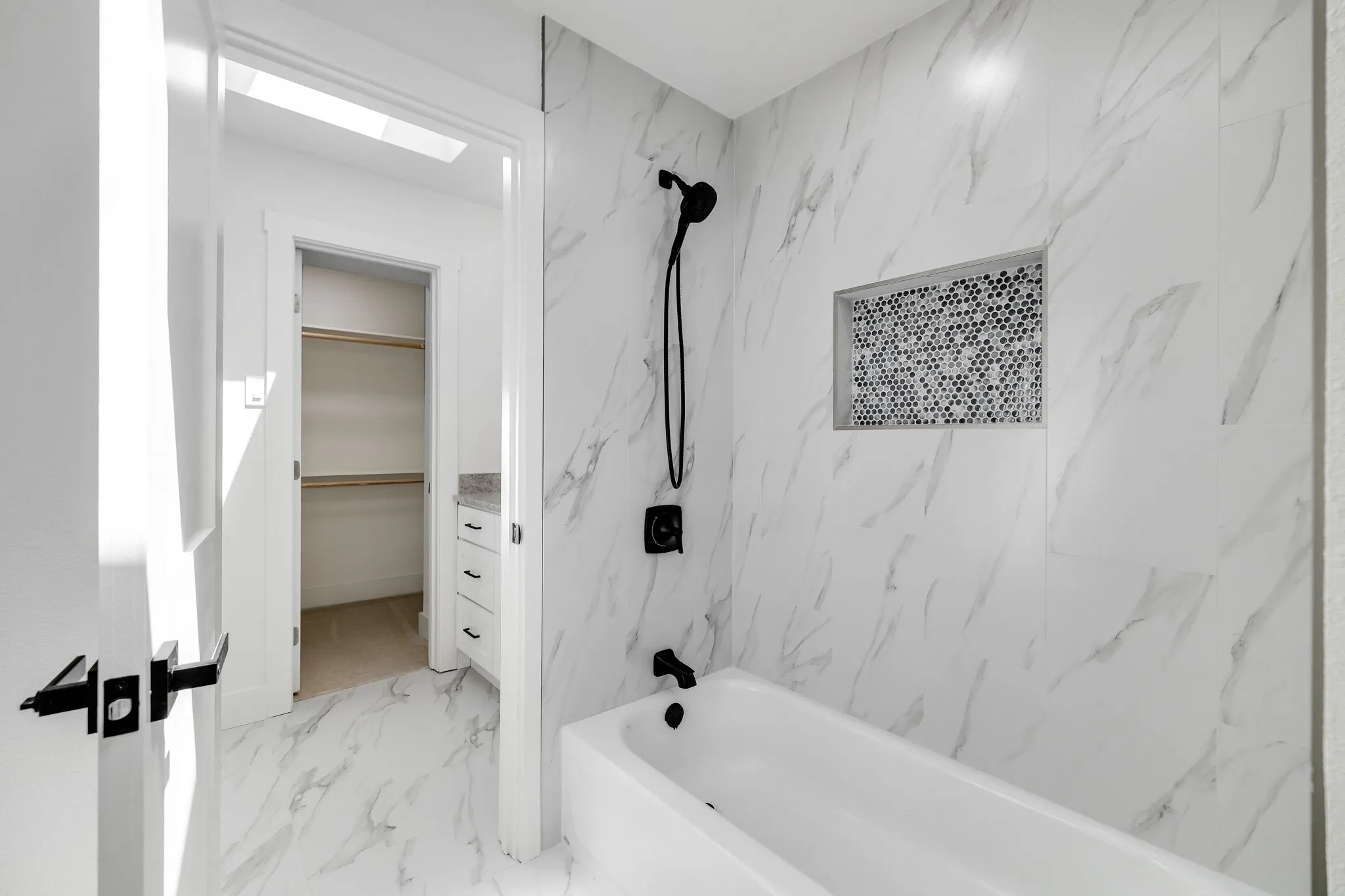 4402 Briargrove-Spa-inspired bathroom featuring marble-look tile walls, a white soaking tub with chrome fixtures and handheld showerhead, a recessed niche with mosaic tile accent, and direct access to a walk-in closet