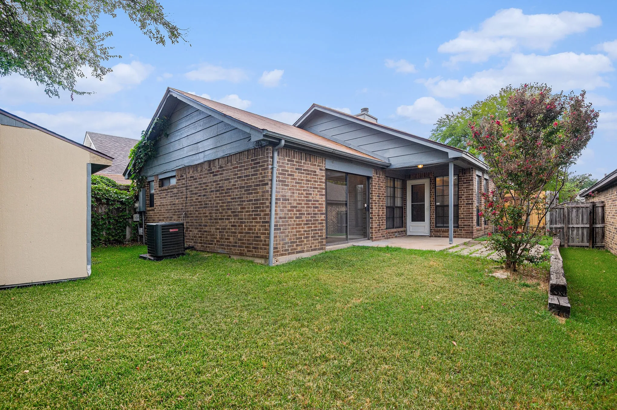 Expansive backyard featuring a manicured lawn, brick exterior, covered patio, and mature landscaping