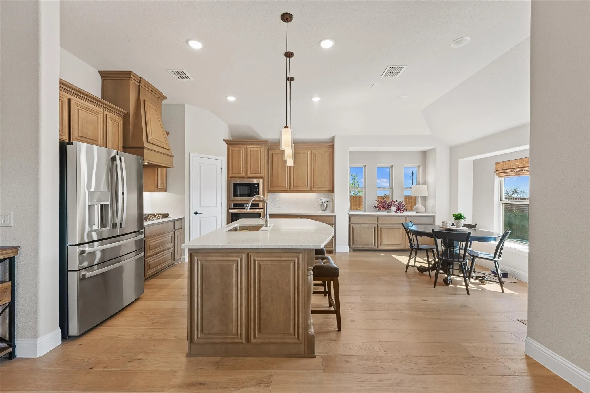 Large kitchen with oversized island, quartz countertops, stainless steel appliances, extensive cabinetry, engineered hardwood floors, pendant lighting, and adjacent breakfast area with large windows.