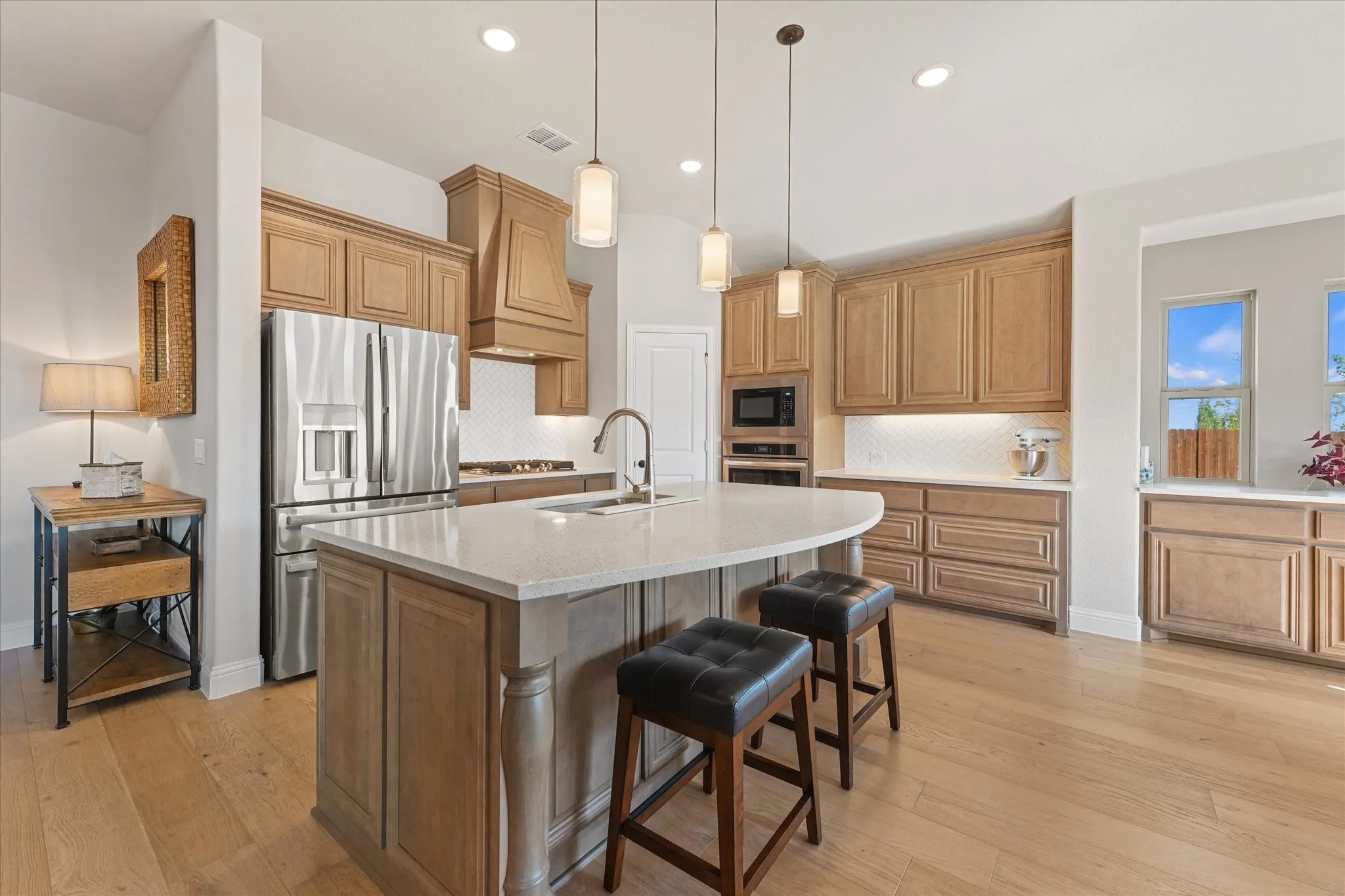 Upgraded kitchen features a large quartz island with seating, stainless steel appliances, built-in oven and microwave, gas cooktop, decorative vent hood, tile backsplash, and extensive cabinet and counter space.