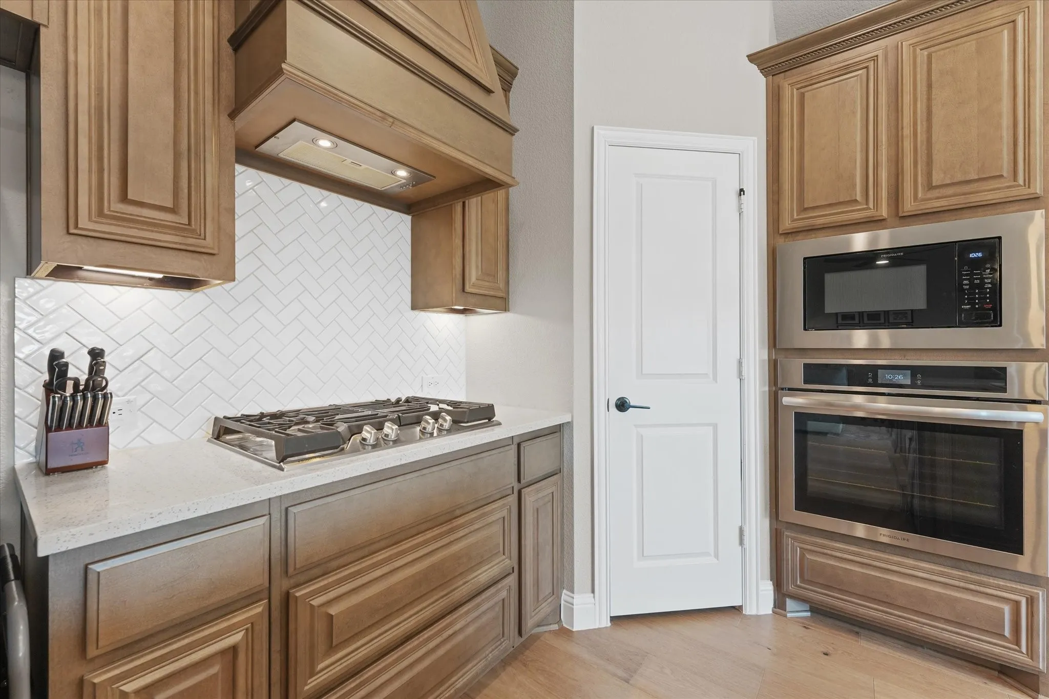 Kitchen includes a gas cooktop with decorative vent hood, herringbone tile backsplash, quartz countertops, built-in stainless steel oven and microwave, and nearby pantry.