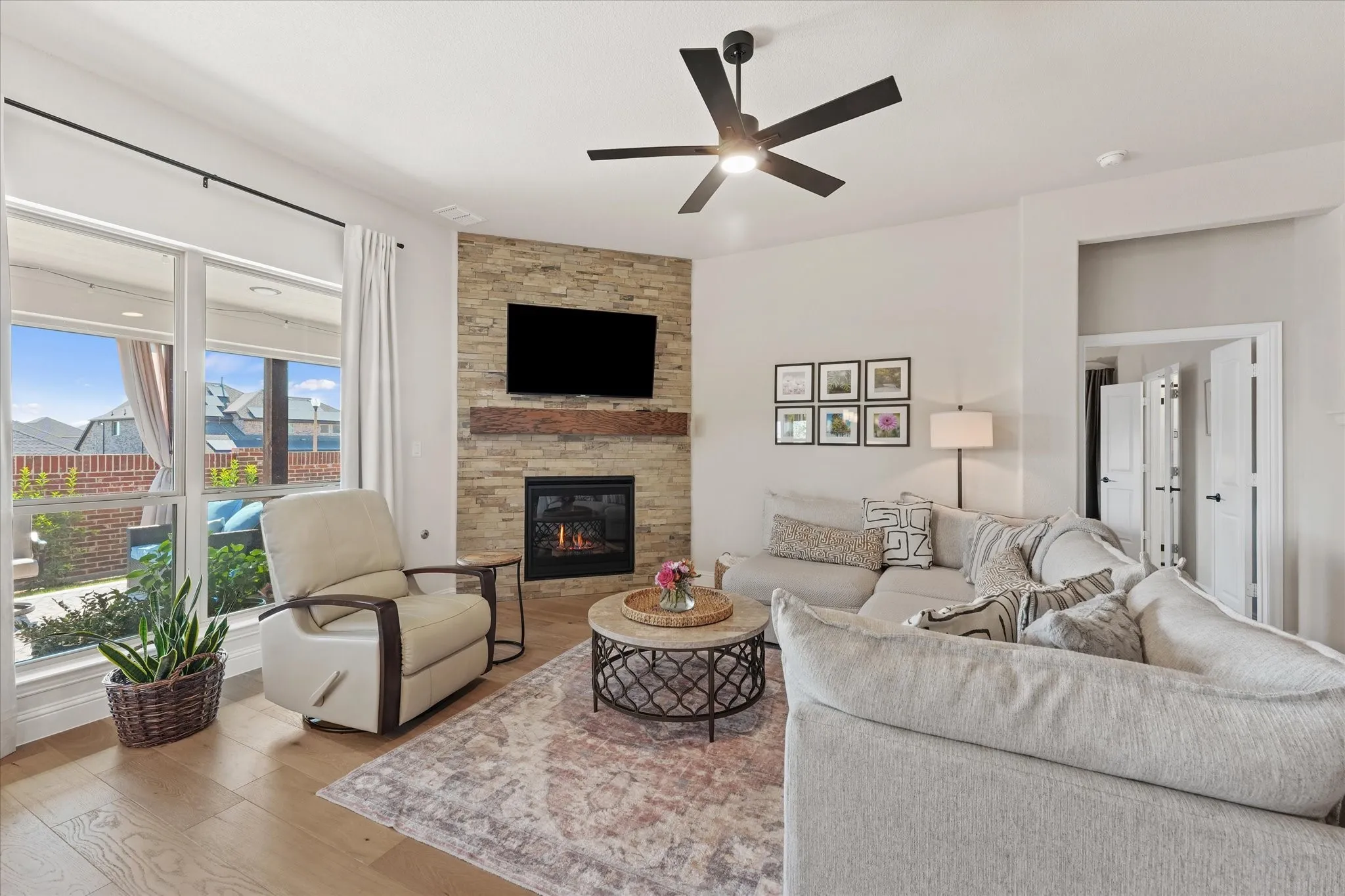 Spacious family room with floor-to-ceiling stone fireplace, wood mantel, large windows, ceiling fan, and engineered hardwood flooring. The open layout connects the living room to the kitchen and dining areas.