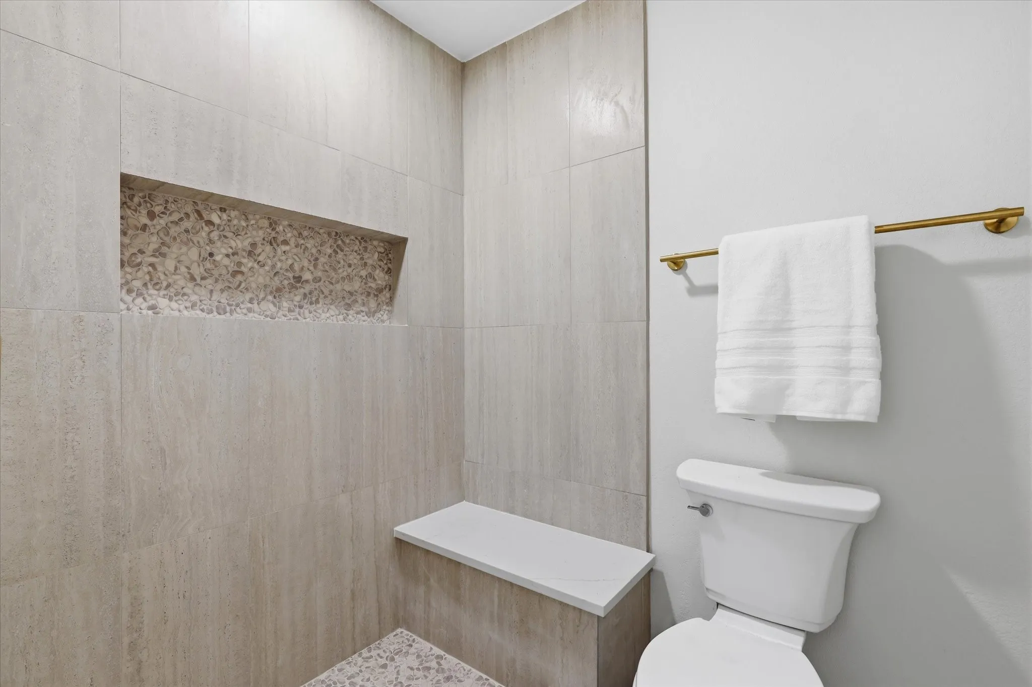 Tiled shower with a built-in niche featuring decorative stone, an integrated shower bench with a solid surface top, and a white toilet with a brass towel bar
