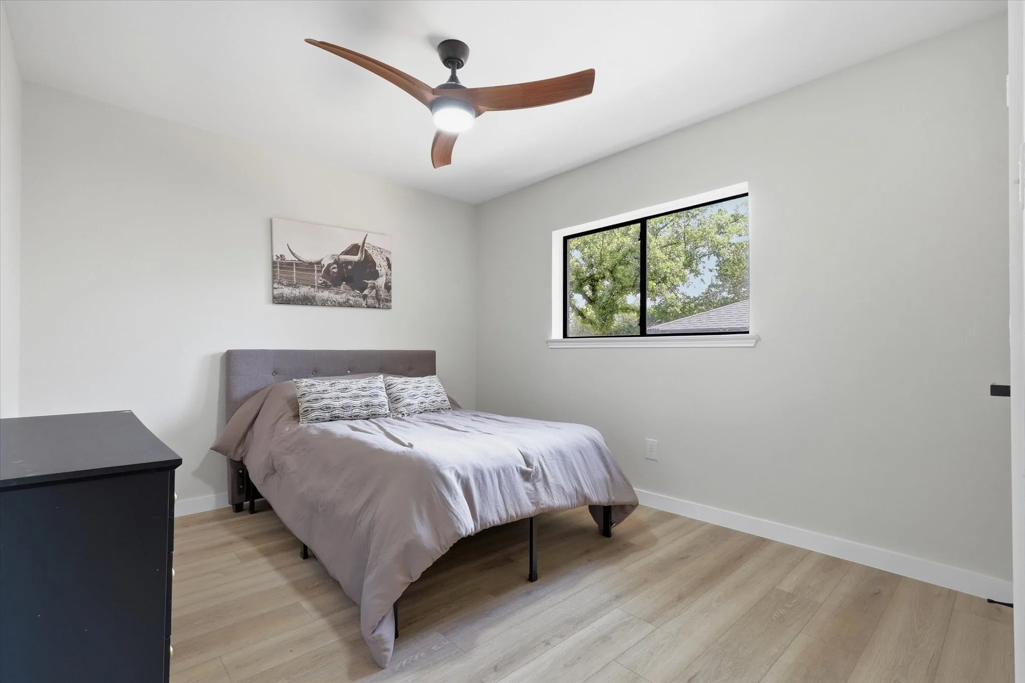 Light wood-finish flooring with crisp white baseboards, a modern ceiling fan with integrated lighting, and a window with black trim