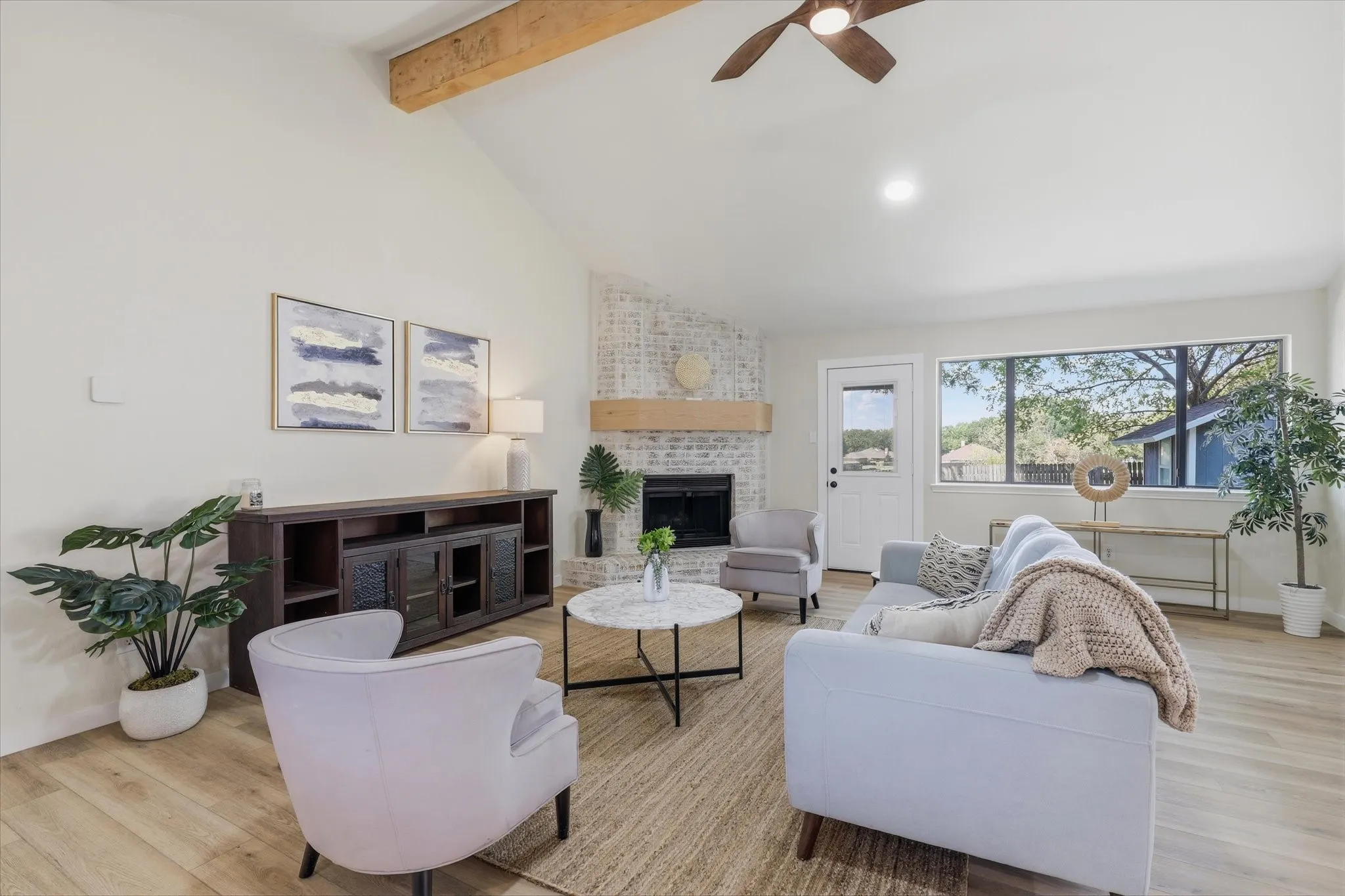 Vaulted ceiling with exposed wood beam, wood-finish flooring, white painted brick fireplace with wood mantel, large window, and ceiling fan
