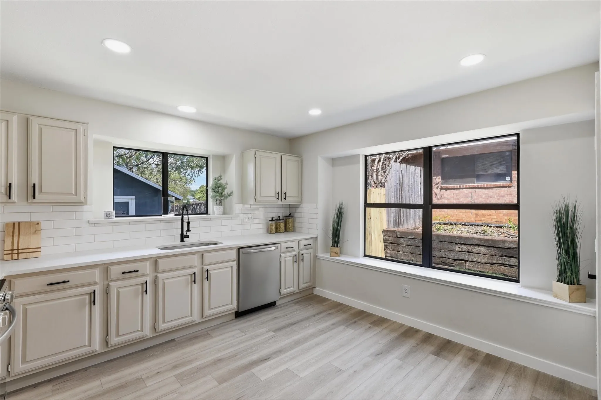 Recessed lighting illuminates the kitchen featuring light-toned cabinetry, a stainless steel dishwasher, white subway tile backsplash, and a large window above the sink