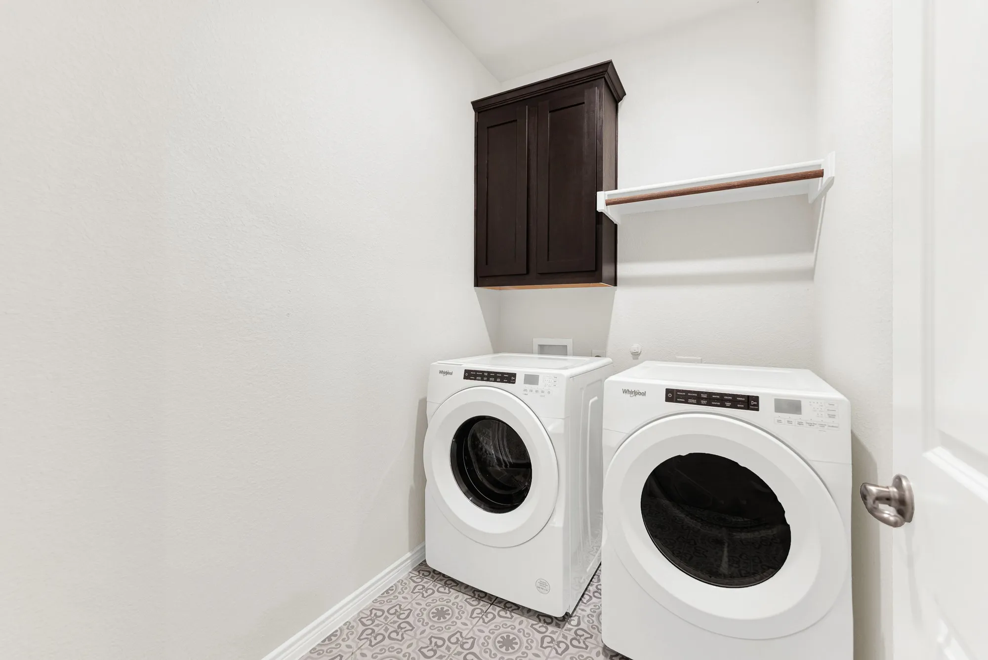 The functional utility room features patterned tile floors, upper cabinetry, and a dedicated hanging rod above modern front-load laundry units. Keep daily chores organized and hassle-free in this clean, stylish, and practical space.