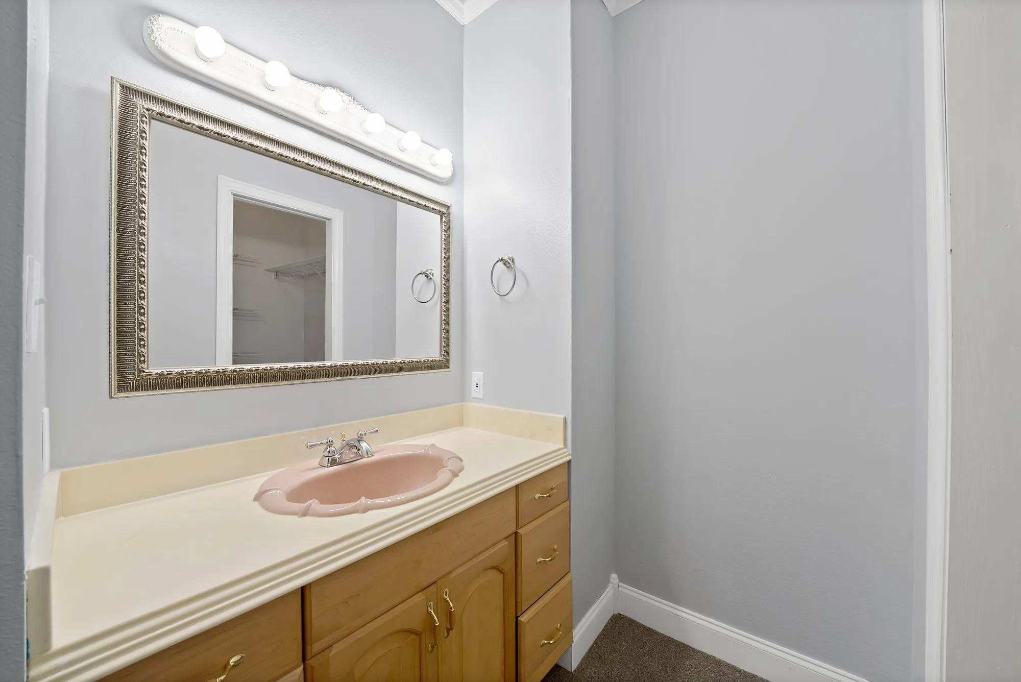 Light wood-finish vanity with gold-tone hardware, a light-colored countertop, and a vanity mirror with an ornate frame. This is an additional sink in the bedroom.