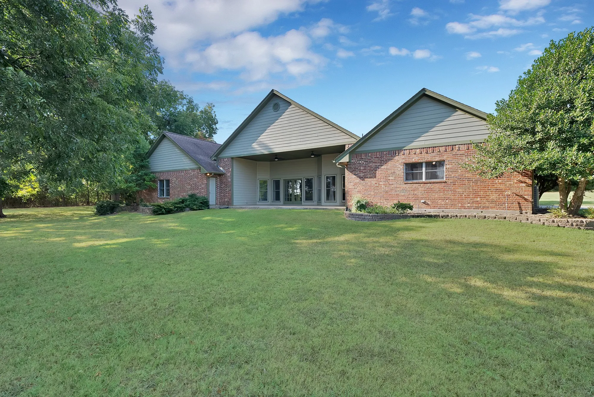 Expansive green lawn with mature trees, brick exterior, and covered patio area with multiple glass doors