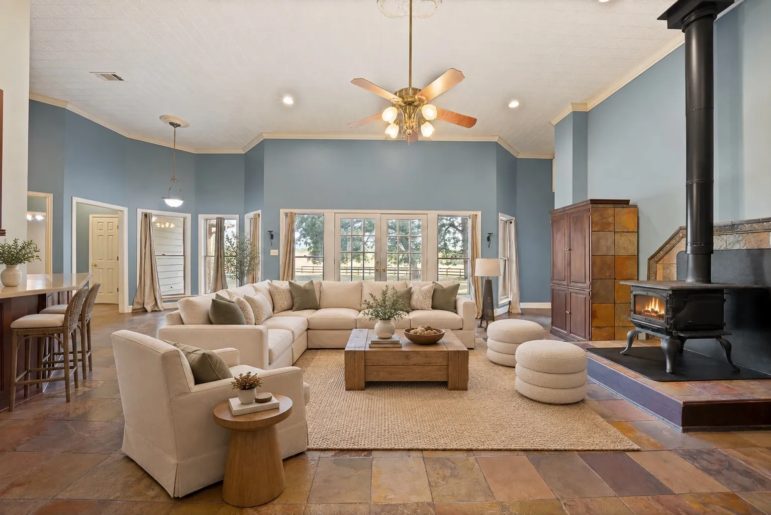 Photo has been virtually staged. Expansive living space featuring high ceilings, a ceiling fan, multiple windows with exterior access, a wood-burning stove with a raised hearth, and natural stone tile flooring