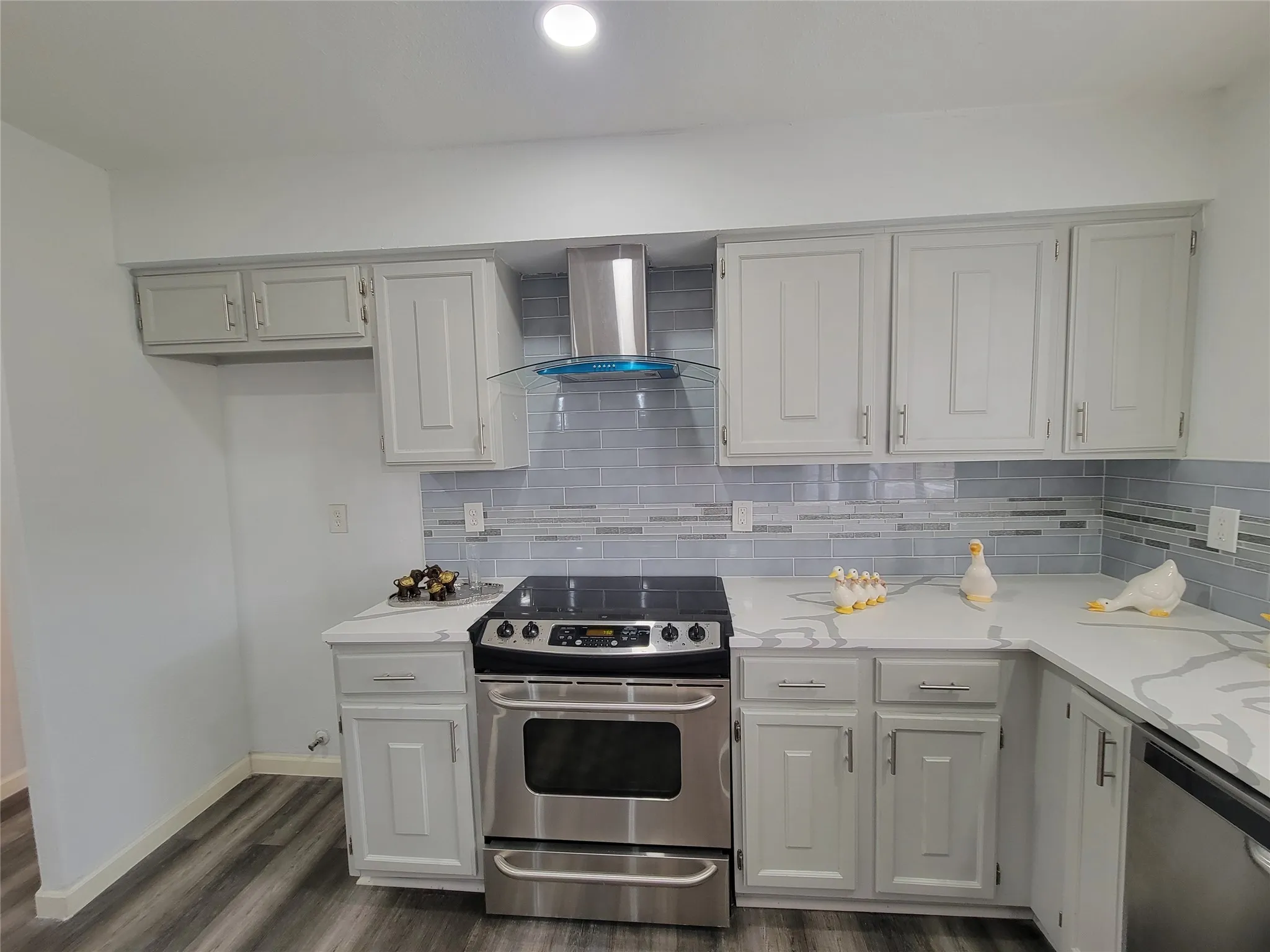 Kitchen featuring dark wood-style flooring, baseboards, decorative backsplash, wall chimney range hood, and appliances with stainless steel finishes