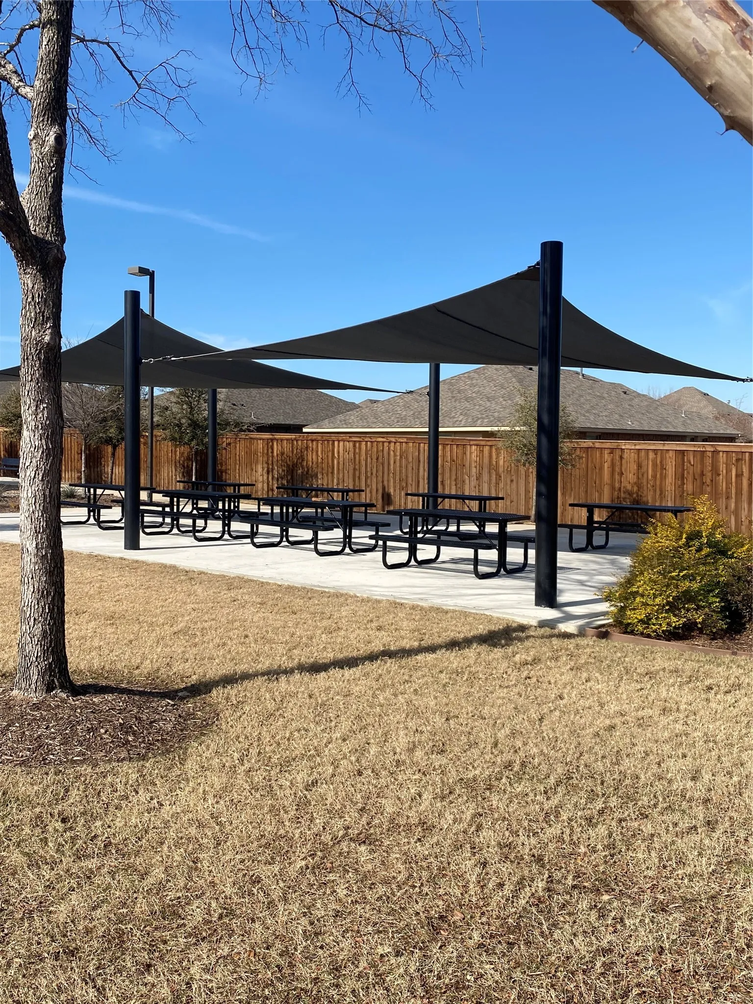 Shaded outdoor seating area featuring multiple picnic tables, concrete pathways, and a perimeter wood fence