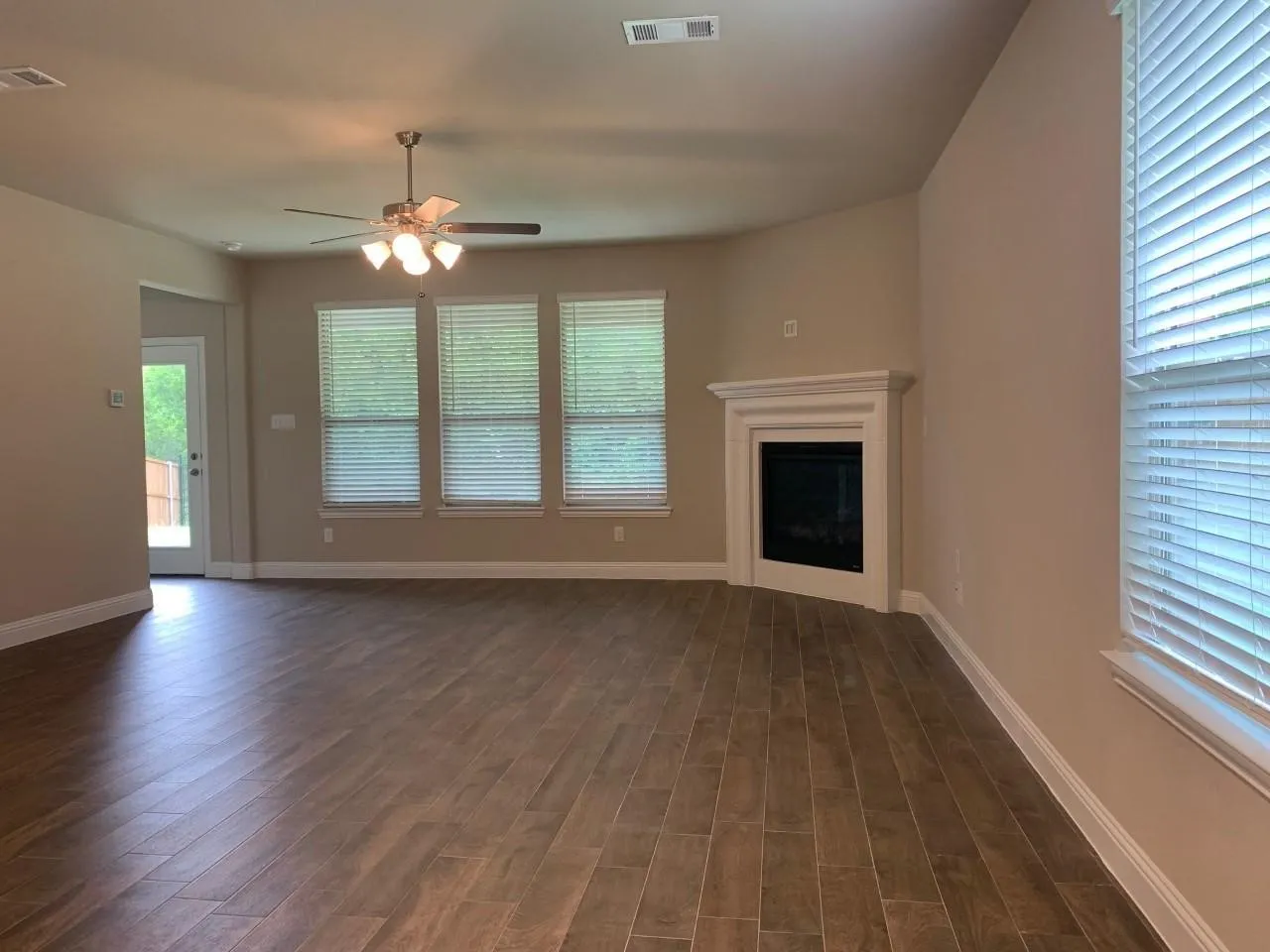 Wood-finish tile flooring throughout, white trim, corner fireplace with white mantel, ceiling fan with light fixture, and multiple windows with blinds