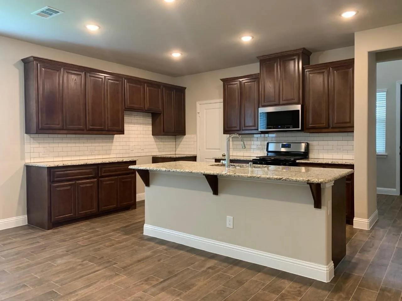 Kitchen featuring granite countertops, dark wood-finish cabinetry, subway tile backsplash, stainless steel appliances, and wood-look tile flooring