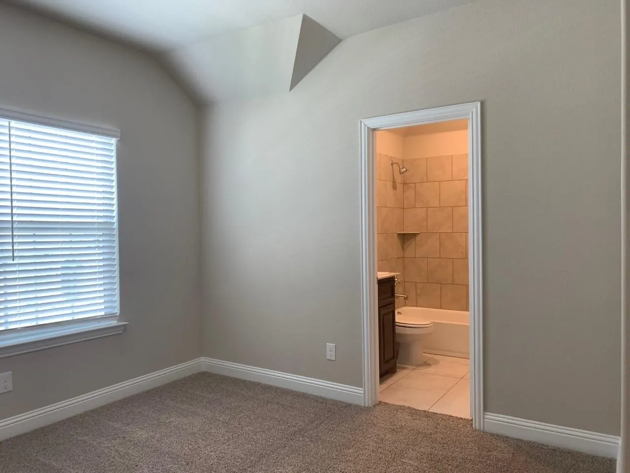 Vaulted ceiling, carpet flooring, window with blinds, bathroom access, and tiled shower surround