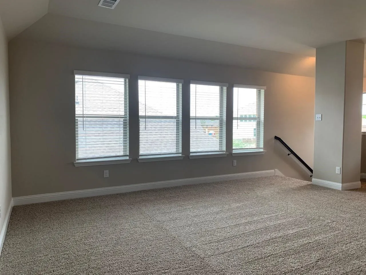 Expansive room featuring four windows with blinds, neutral carpeting, and a black handrail