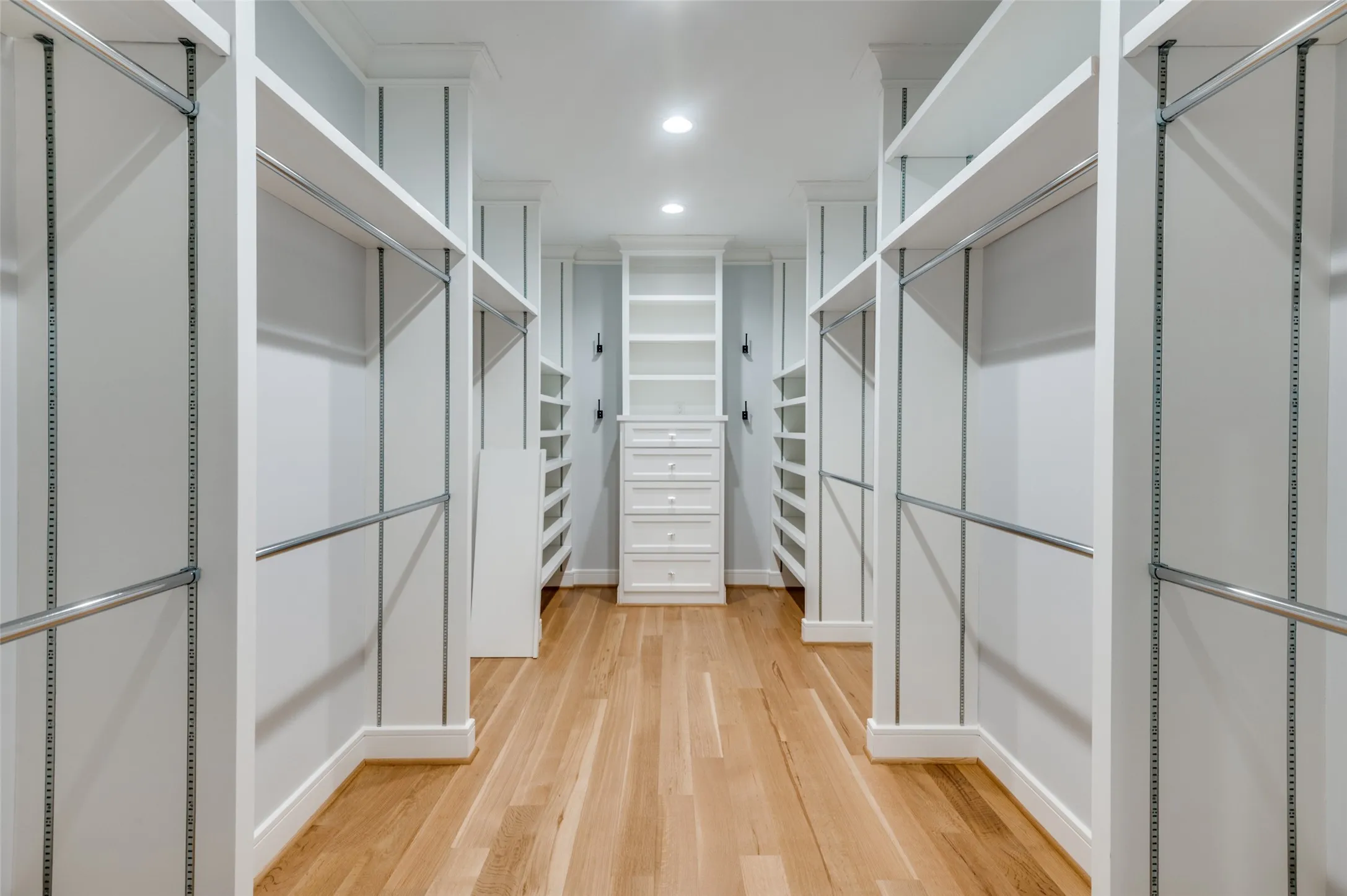 Walk-in closet with natural wood-finish flooring, recessed lighting, white built-in shelving, chrome hanging rods, and a central dresser with multiple drawers
