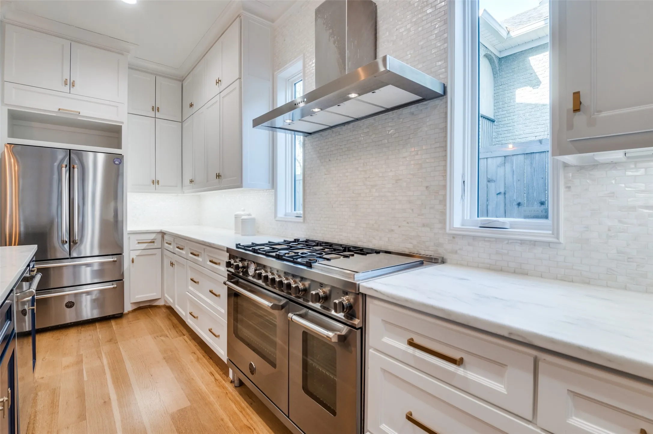 White cabinetry with gold-tone hardware, stainless steel appliances, wood-finish flooring, and a white tile backsplash