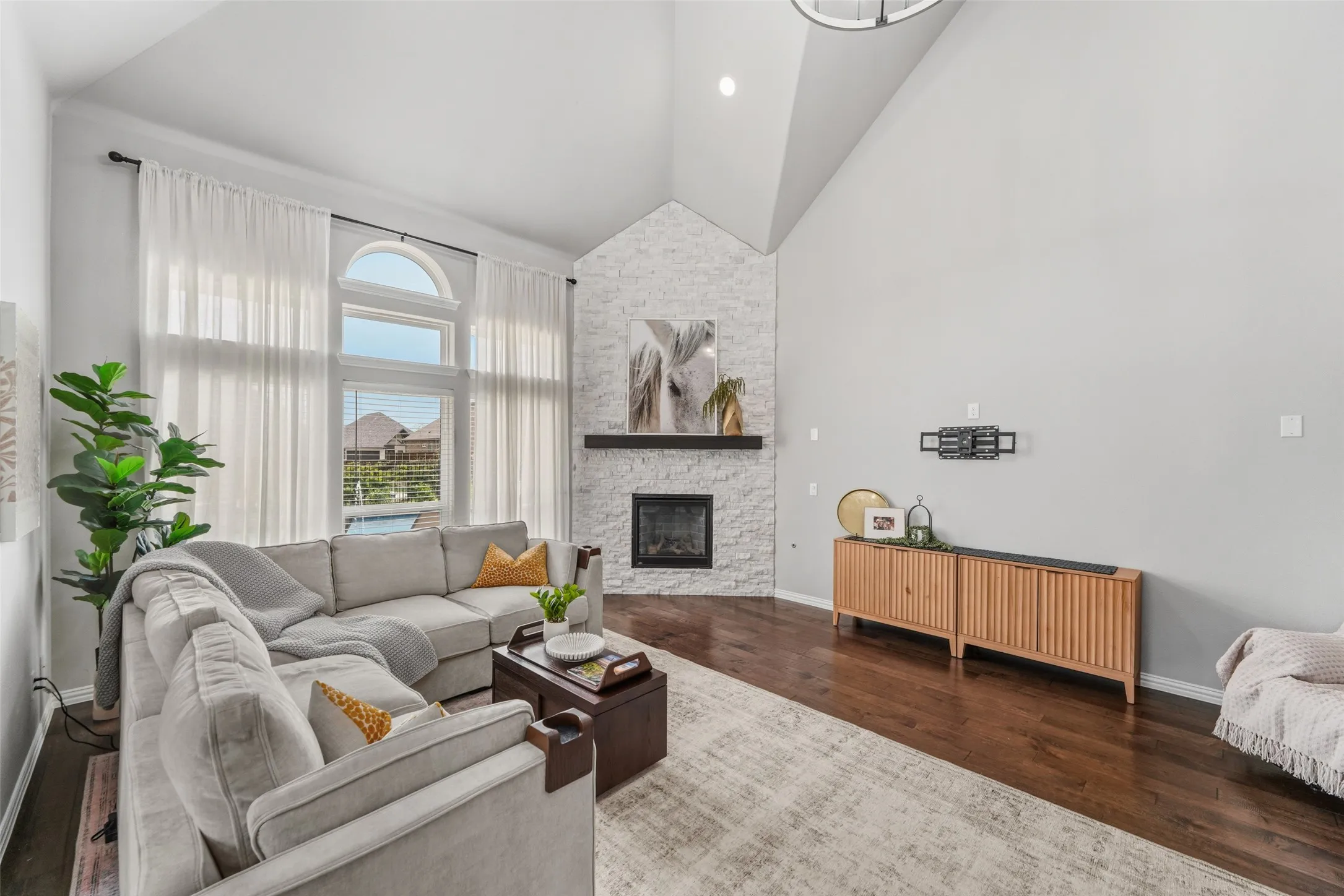 Vaulted ceiling, corner fireplace with stone surround, dark wood-finish flooring, and large windows with transom
