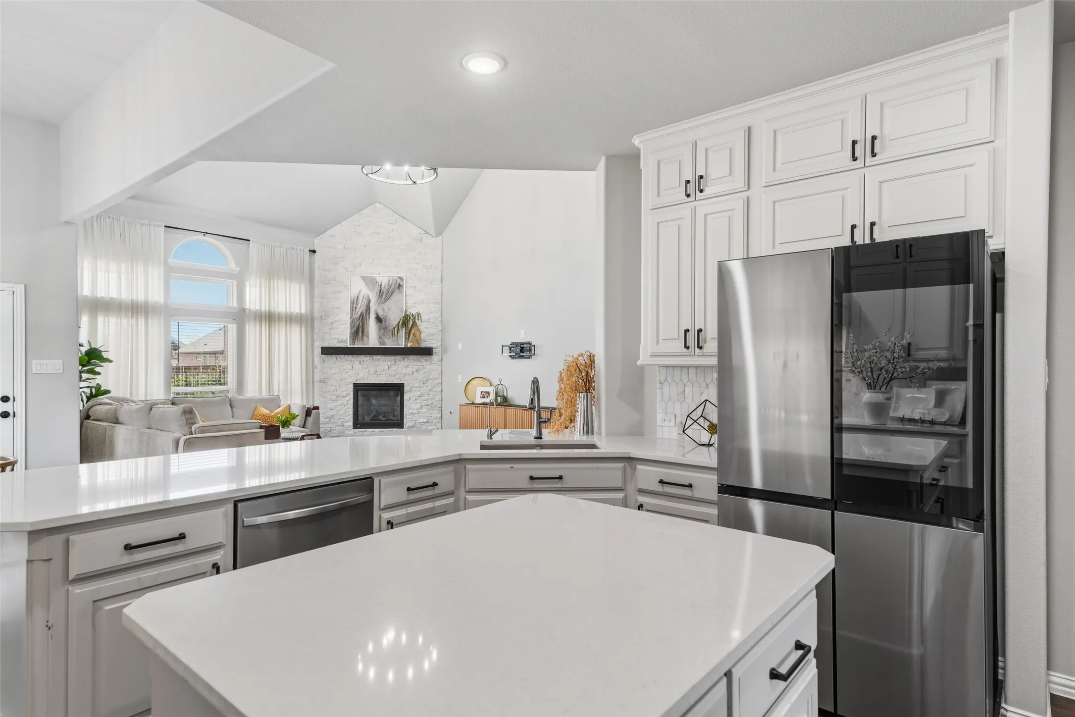 White cabinetry with black hardware, stainless steel appliances, light-colored countertops, recessed lighting, and a stone fireplace
