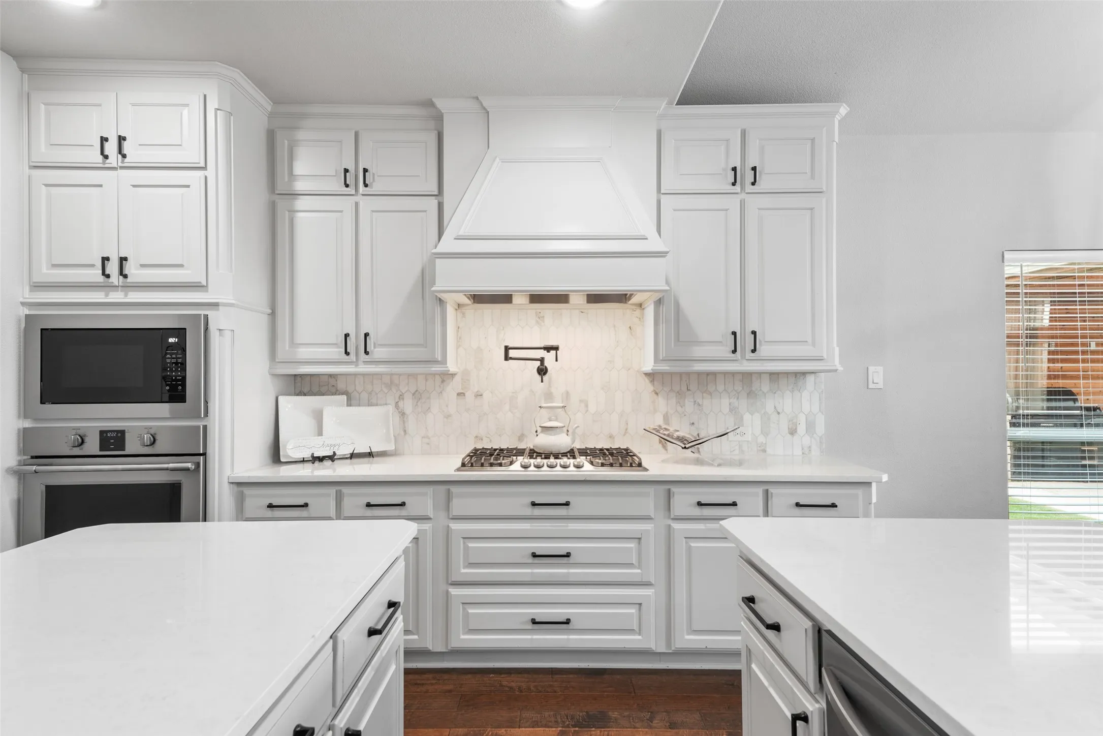 White shaker-style cabinetry, stainless steel built-in microwave and oven, gas cooktop with pot filler, custom range hood, and light-toned countertops