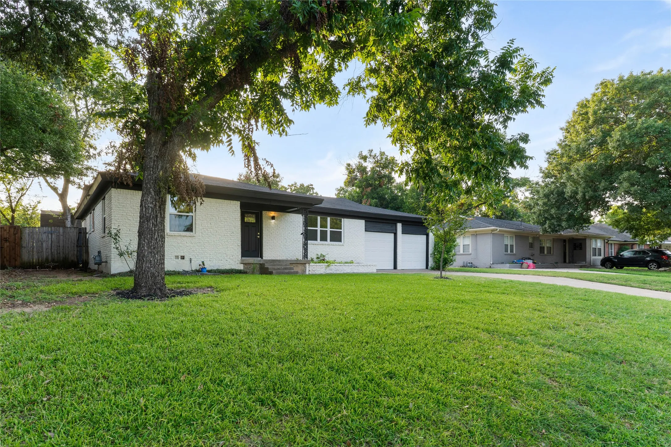 White painted brick exterior with a dark trim roofline, attached two-car garage, mature front yard trees, and a concrete driveway