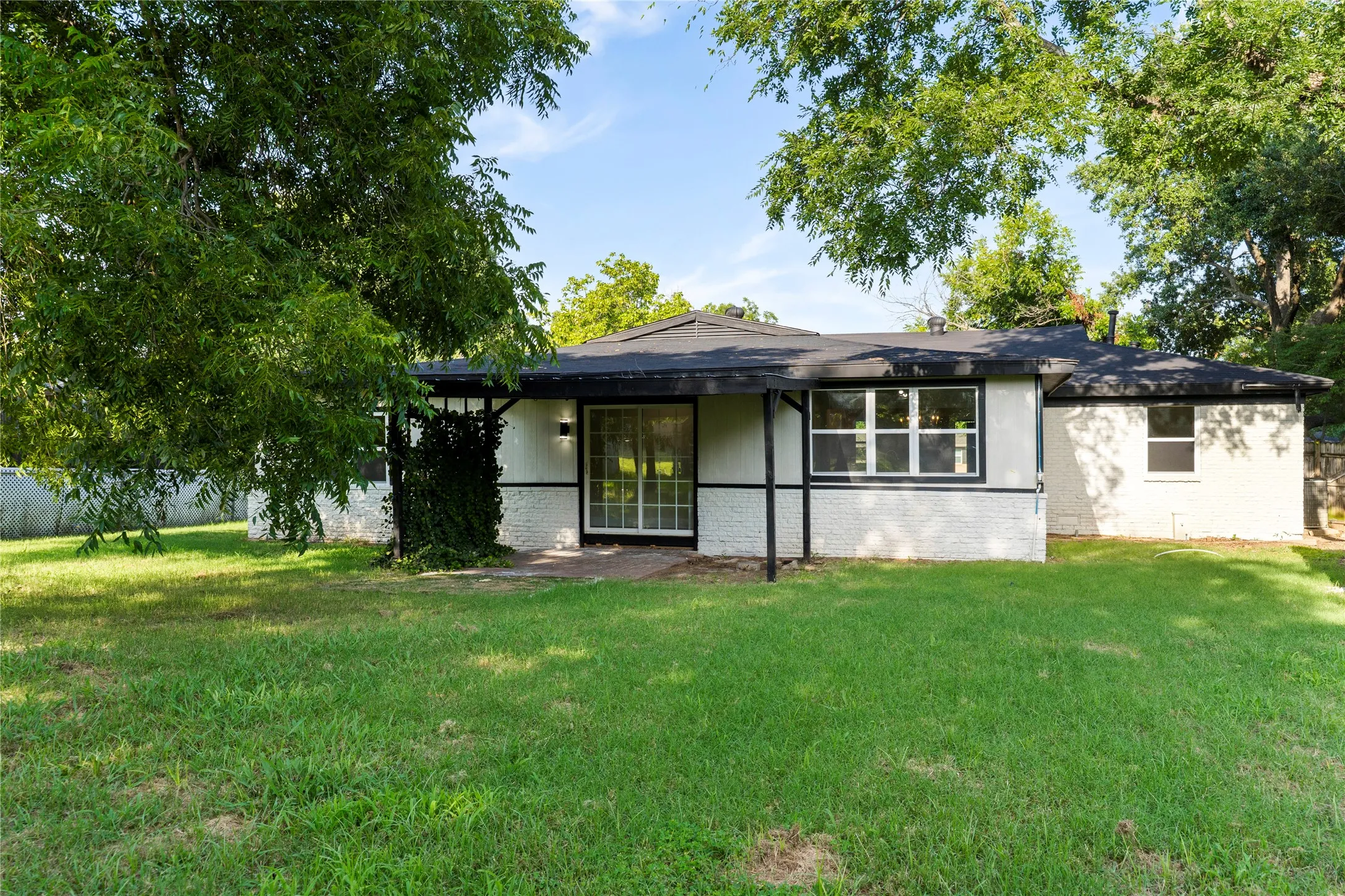 Expansive backyard with mature trees, a covered patio, white brick exterior, and a sliding glass door