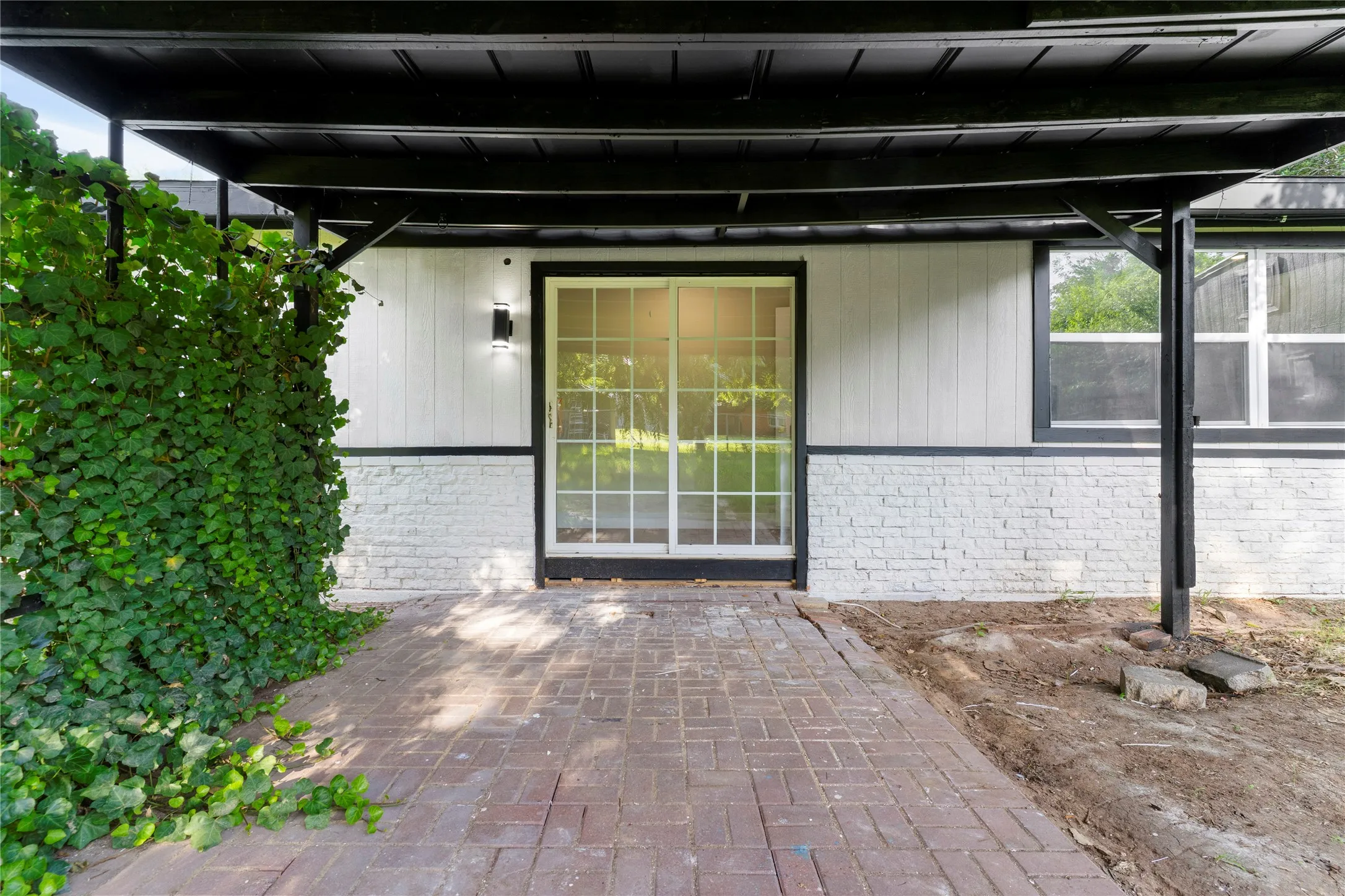 Brick paver patio with an overhead awning, sliding glass door entry, white brick facade, and vertical panel siding