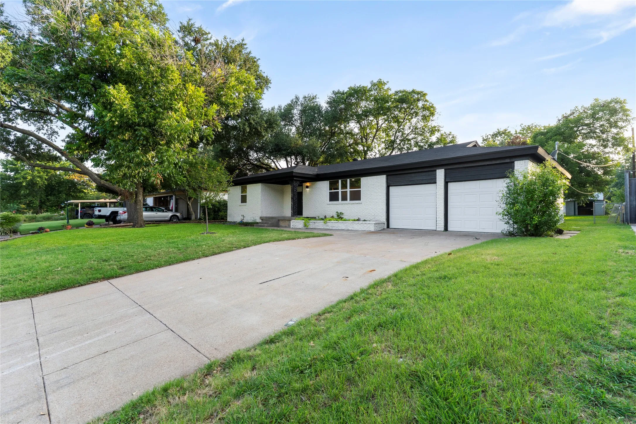 Brick exterior with black trim, a two-car garage, mature trees, a well-maintained lawn, and a concrete driveway