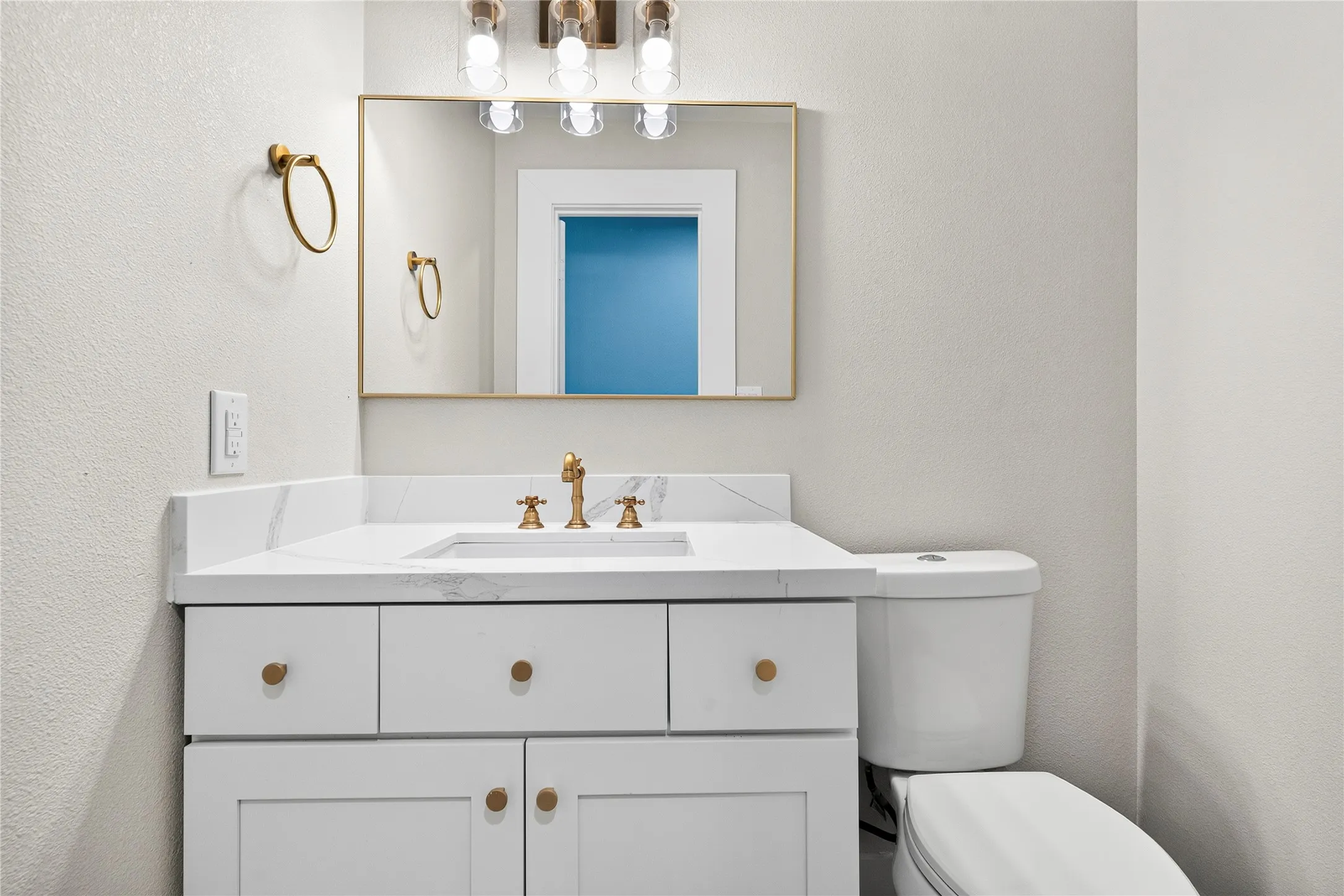White vanity with gold-tone hardware, white countertop, rectangular undermount sink, and a gold-framed mirror