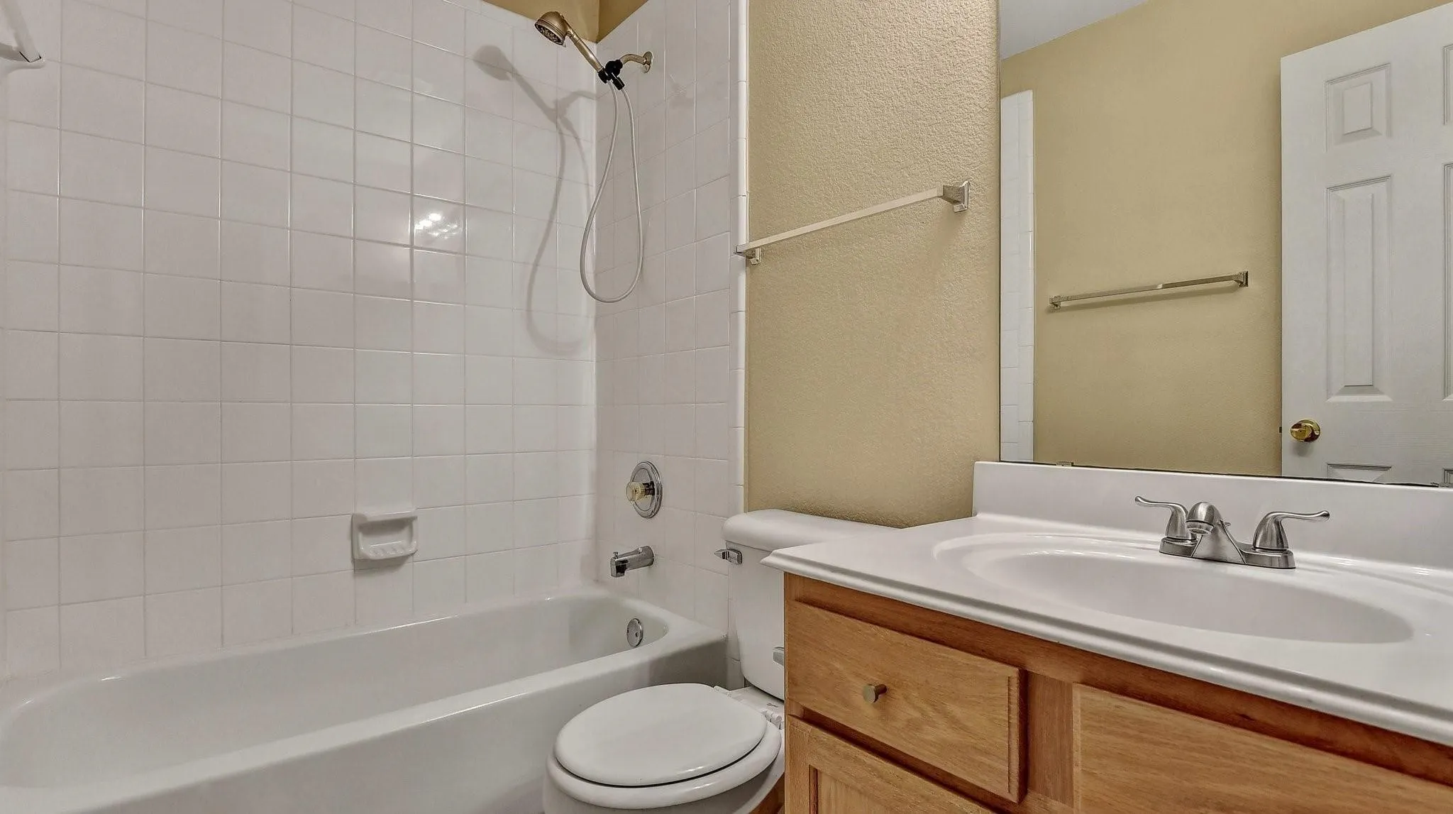 White tiled tub and shower combination with a gold-tone showerhead, wood-finish vanity with a built-in sink, and a large mirror