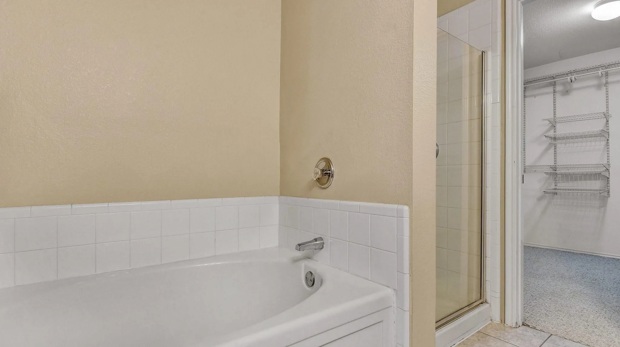Built-in soaking tub with tile surround, enclosed shower, and a separate room featuring built-in wire shelving