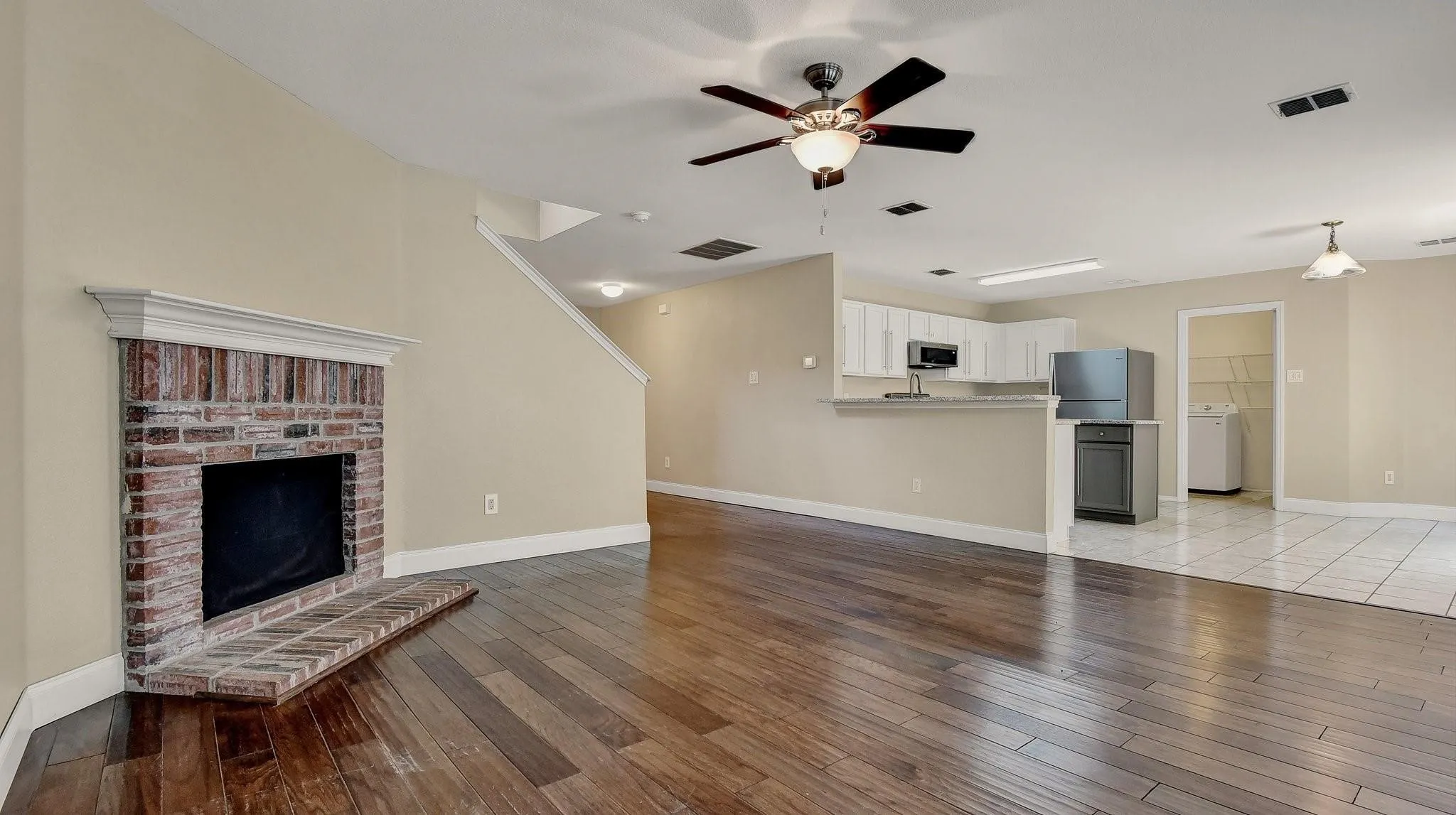 Wood-finish flooring, brick fireplace with mantel, white kitchen cabinetry, kitchen peninsula with counter seating, and ceiling fan with integrated lighting