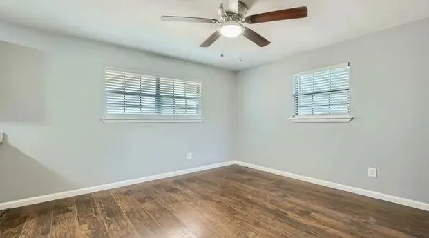 Wood-finish flooring, two windows with blinds, light gray wall paint, and a ceiling fan with integrated lighting