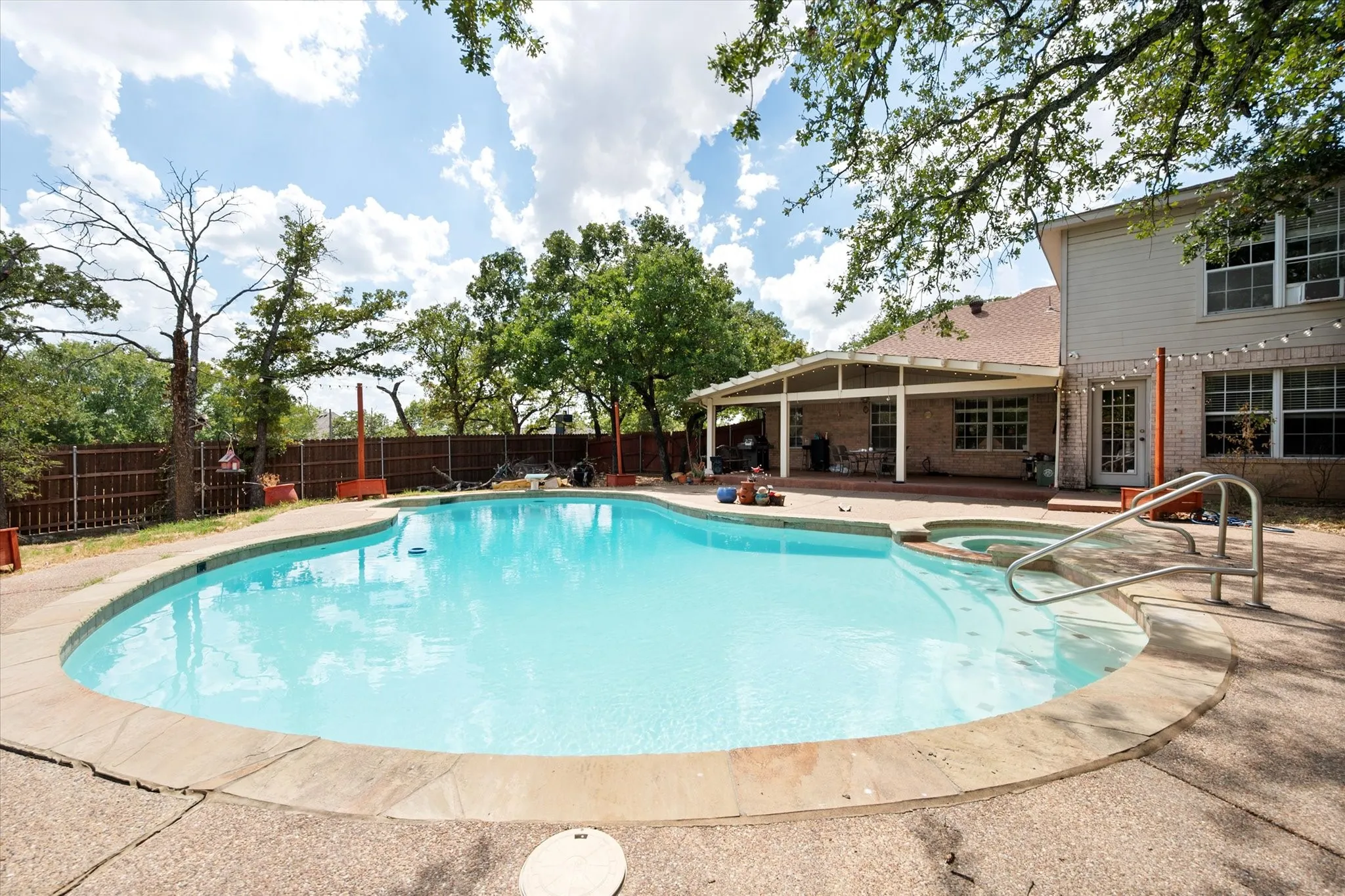 Shaped swimming pool with an integrated spa, expansive paver patio, covered outdoor living area, and a privacy fence