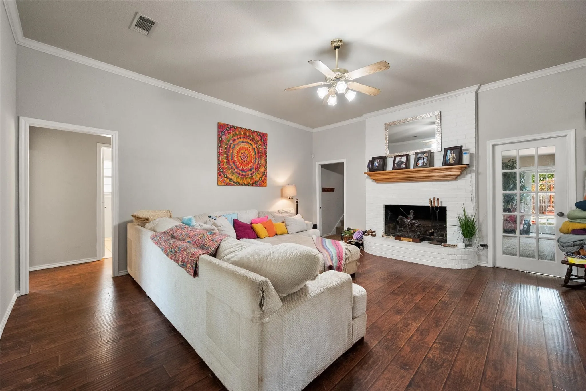 Wood-finish flooring, white brick fireplace with wood mantel, ceiling fan with integrated lighting, and crown molding