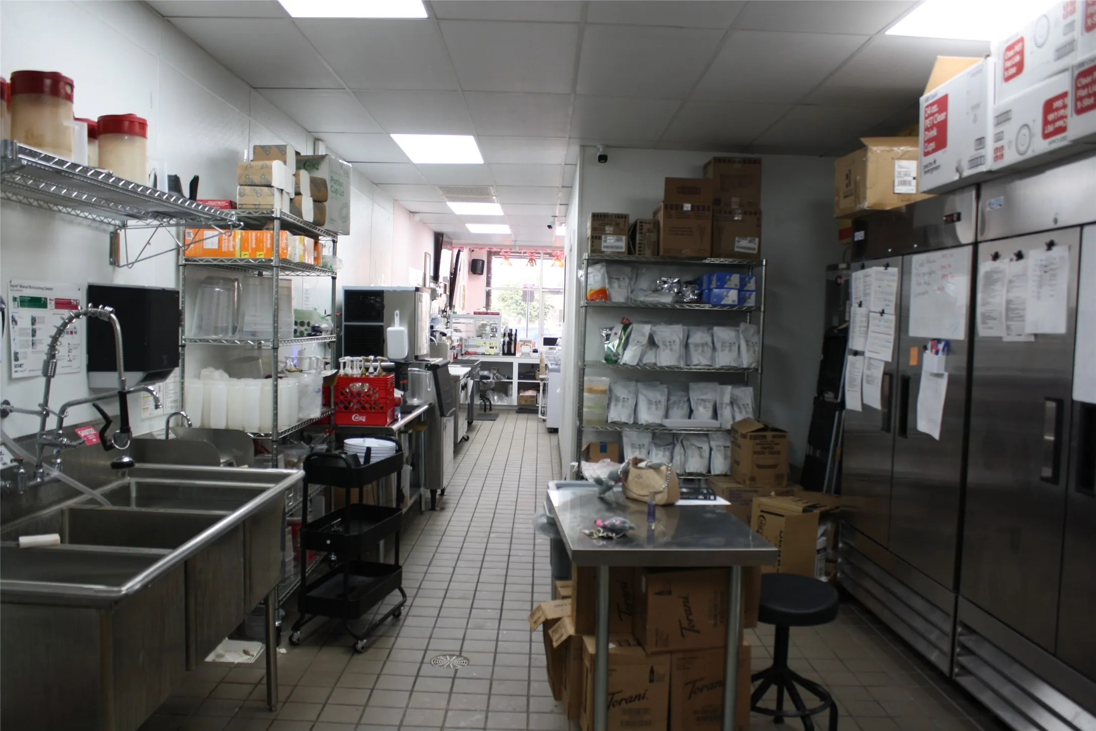 Commercial kitchen featuring stainless steel three-compartment sink, stainless steel shelving, stainless steel refrigeration units, and light-colored tile flooring