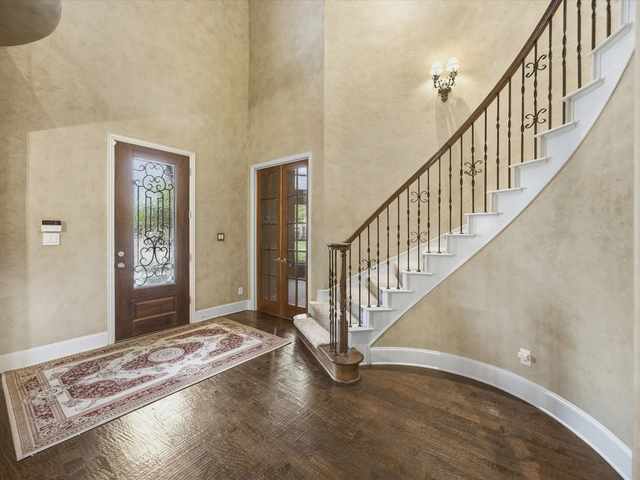 Dramatic two-story entry with wood-finish flooring, a custom front door with decorative ironwork, and a curved staircase featuring wrought iron spindles and a wood handrail
