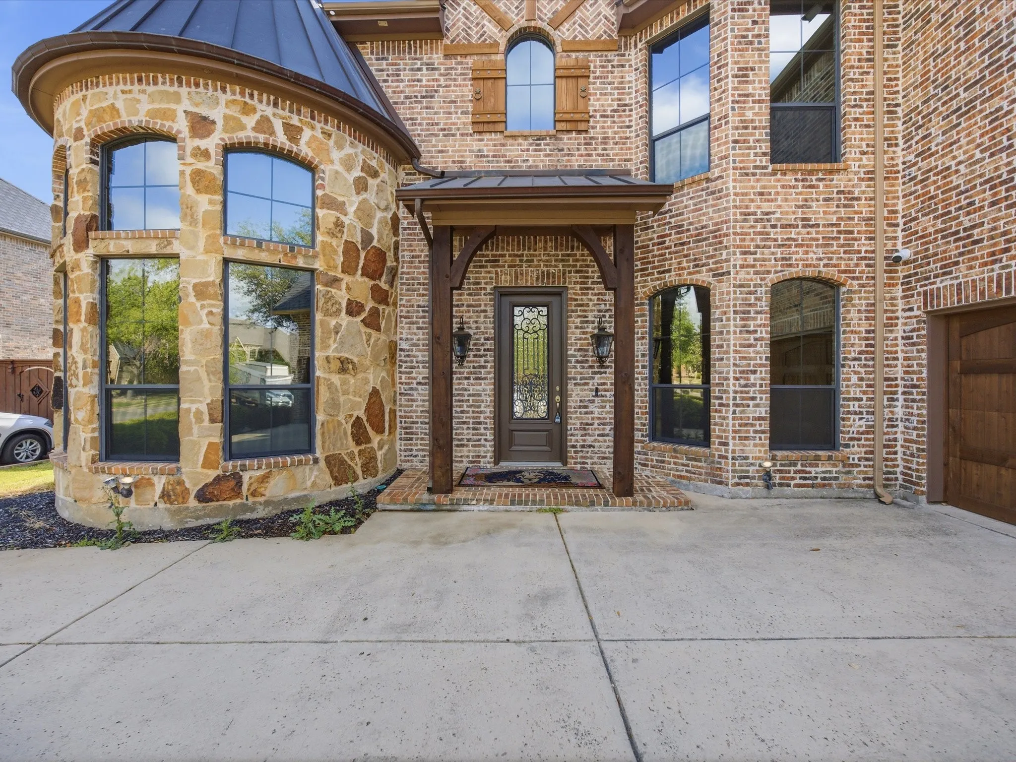 Brick and stone exterior featuring an arched wood-finish entry door with decorative glass, a covered porch with timber supports, and multiple arched and rectangular windows