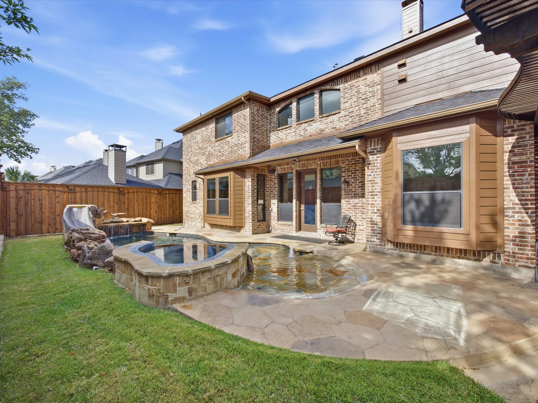 Brick exterior with architectural details, ground-level patio with stone pavers, built-in water feature with rock accents, manicured lawn, and wood privacy fencing