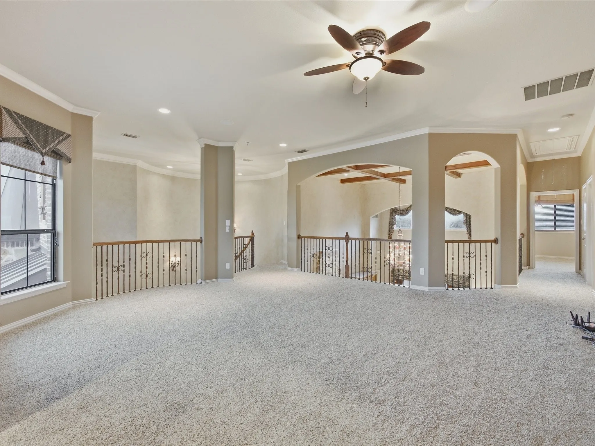 Expansive carpeted loft with a ceiling fan, decorative wrought iron railings, arched openings, and crown molding