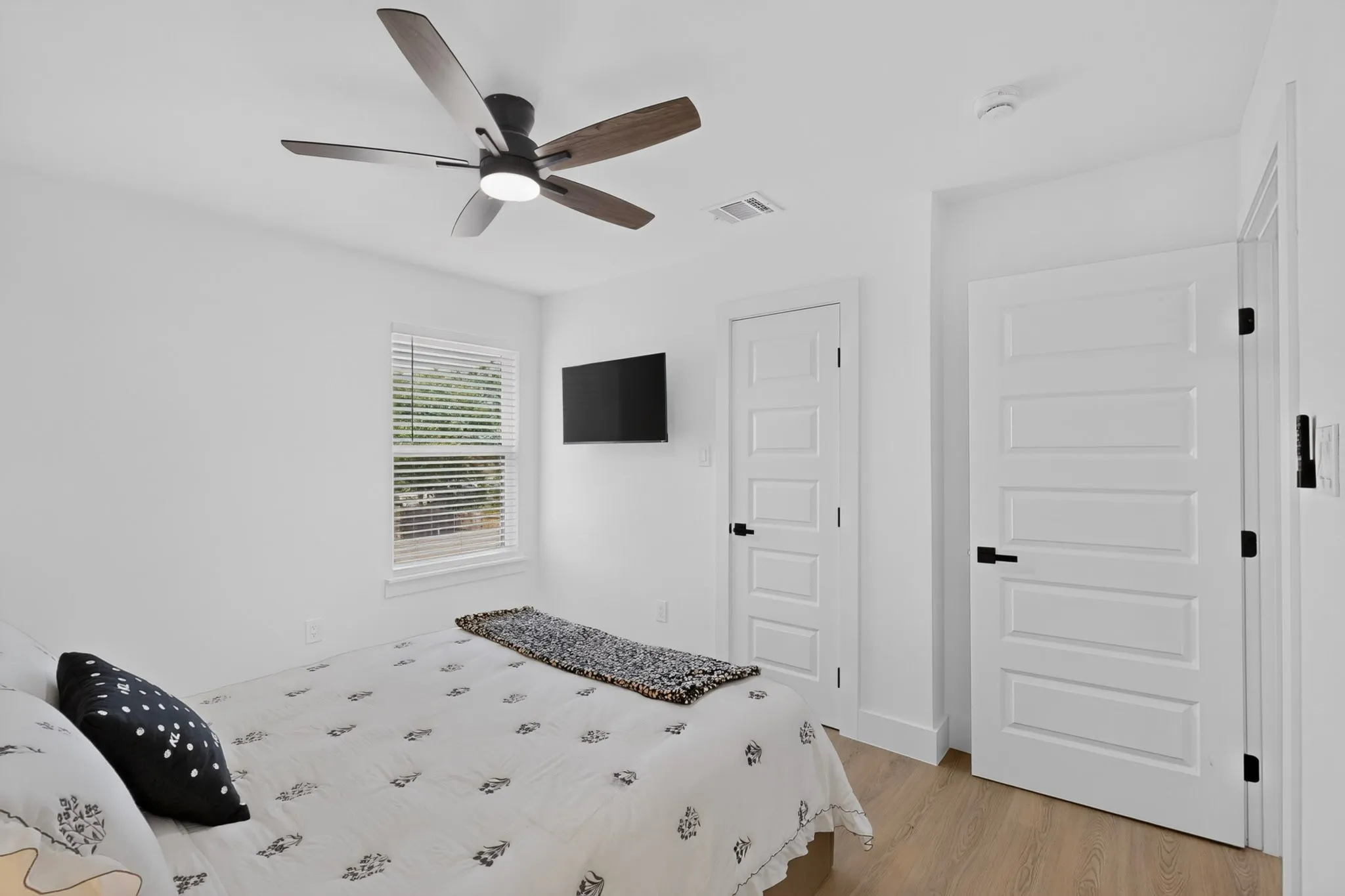 Wood-finish flooring, contemporary ceiling fan with integrated lighting, window with horizontal blinds, and white paneled doors with matte black hardware