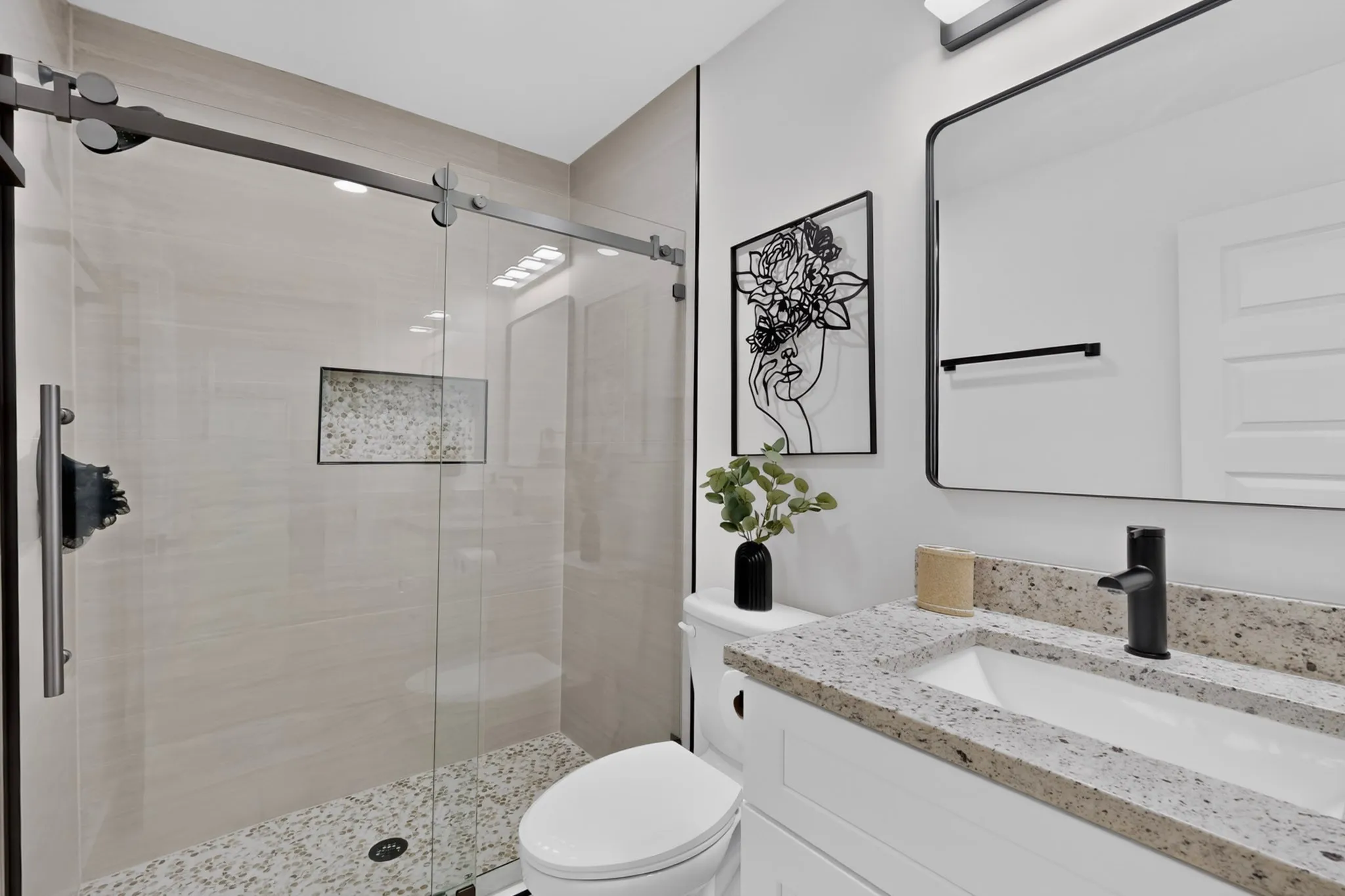 Frameless glass shower enclosure with sliding door, tiled shower surround, recessed shower niche, white vanity with stone-finish countertop, and undermount sink with matte black faucet