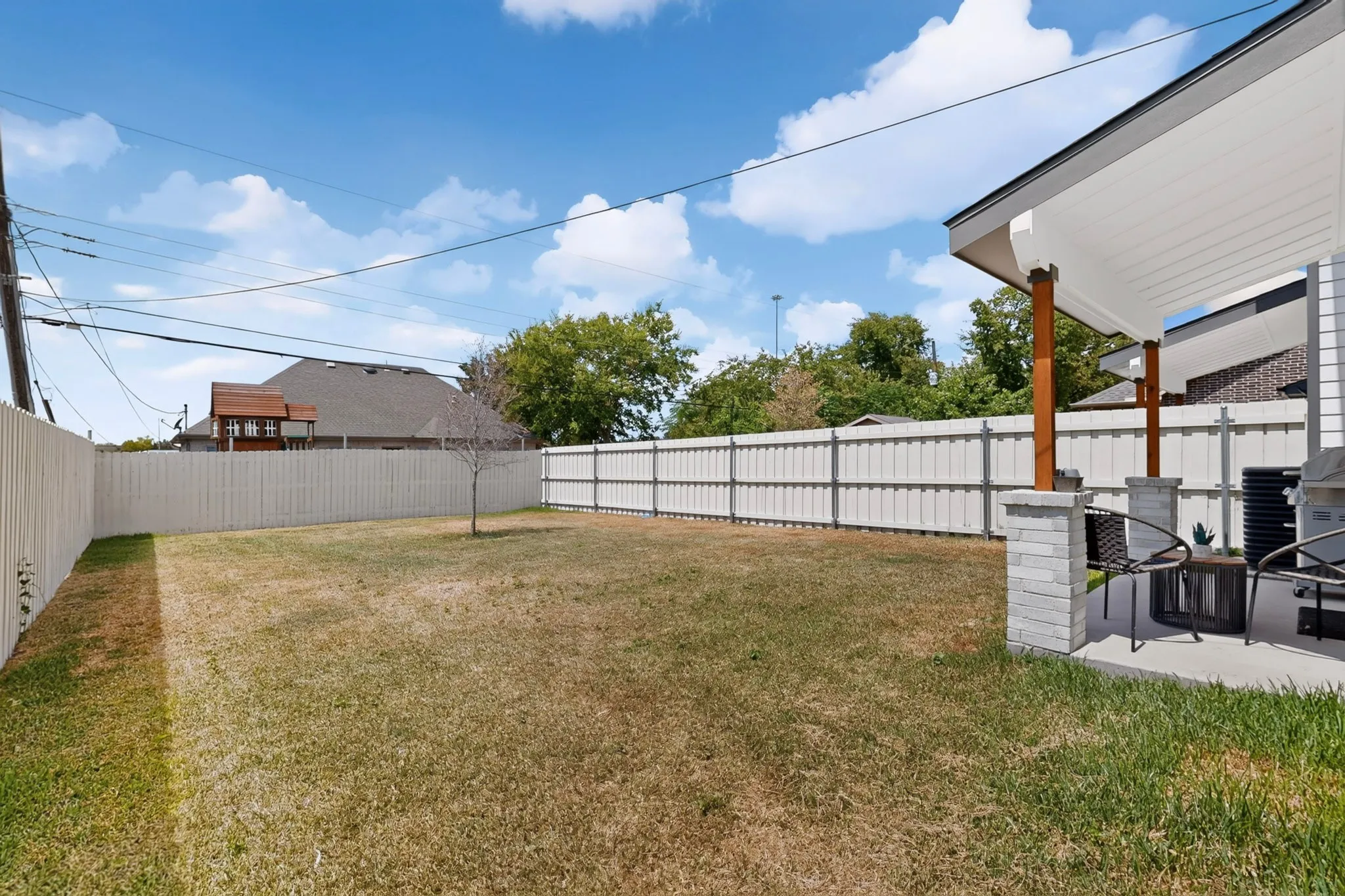 Expansive backyard featuring a concrete patio, white privacy fencing, and a covered porch with stone-clad pillars and wood-tone supports