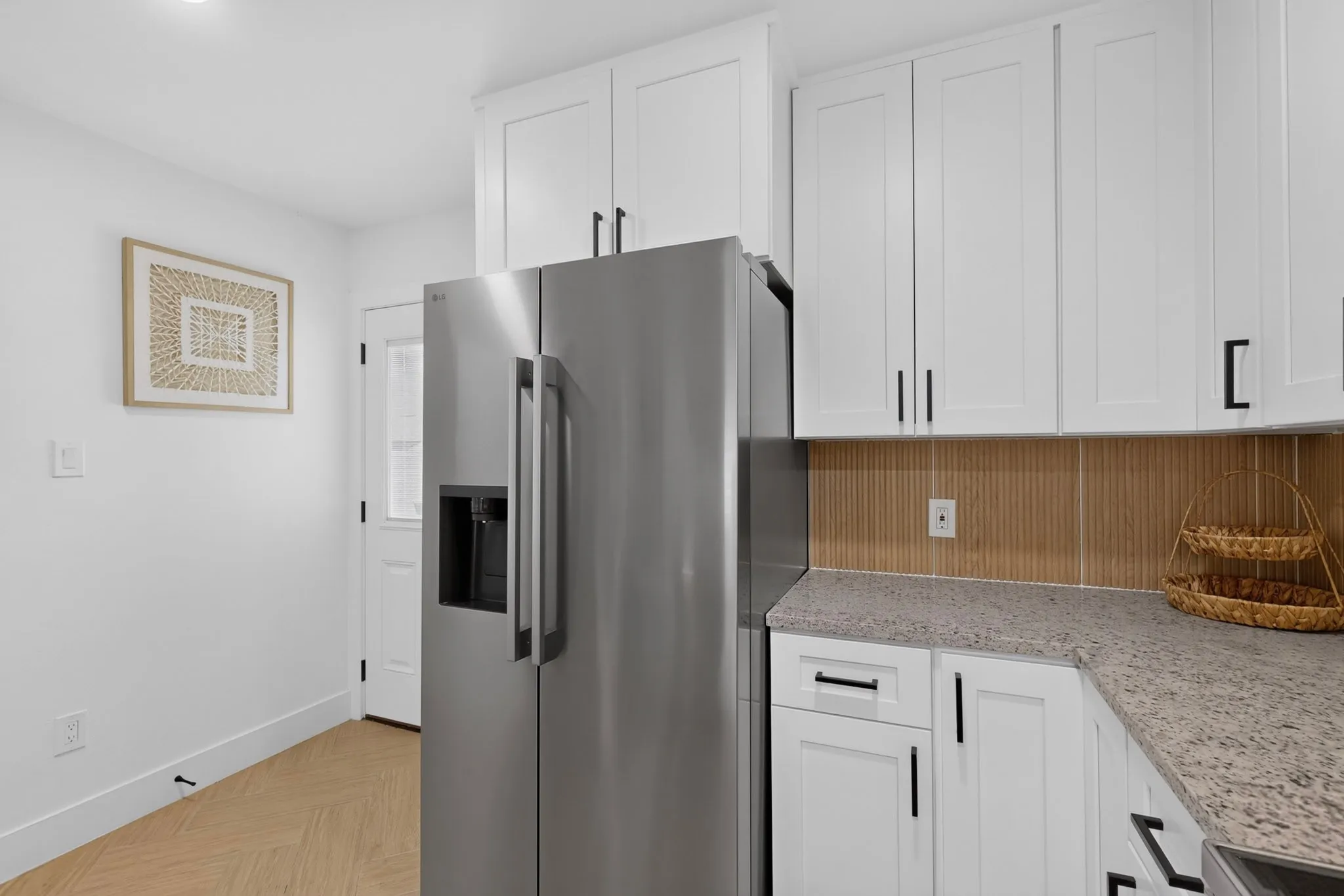 White shaker-style cabinetry with matte black hardware, light-toned countertop, stainless steel refrigerator, and wood-finish flooring