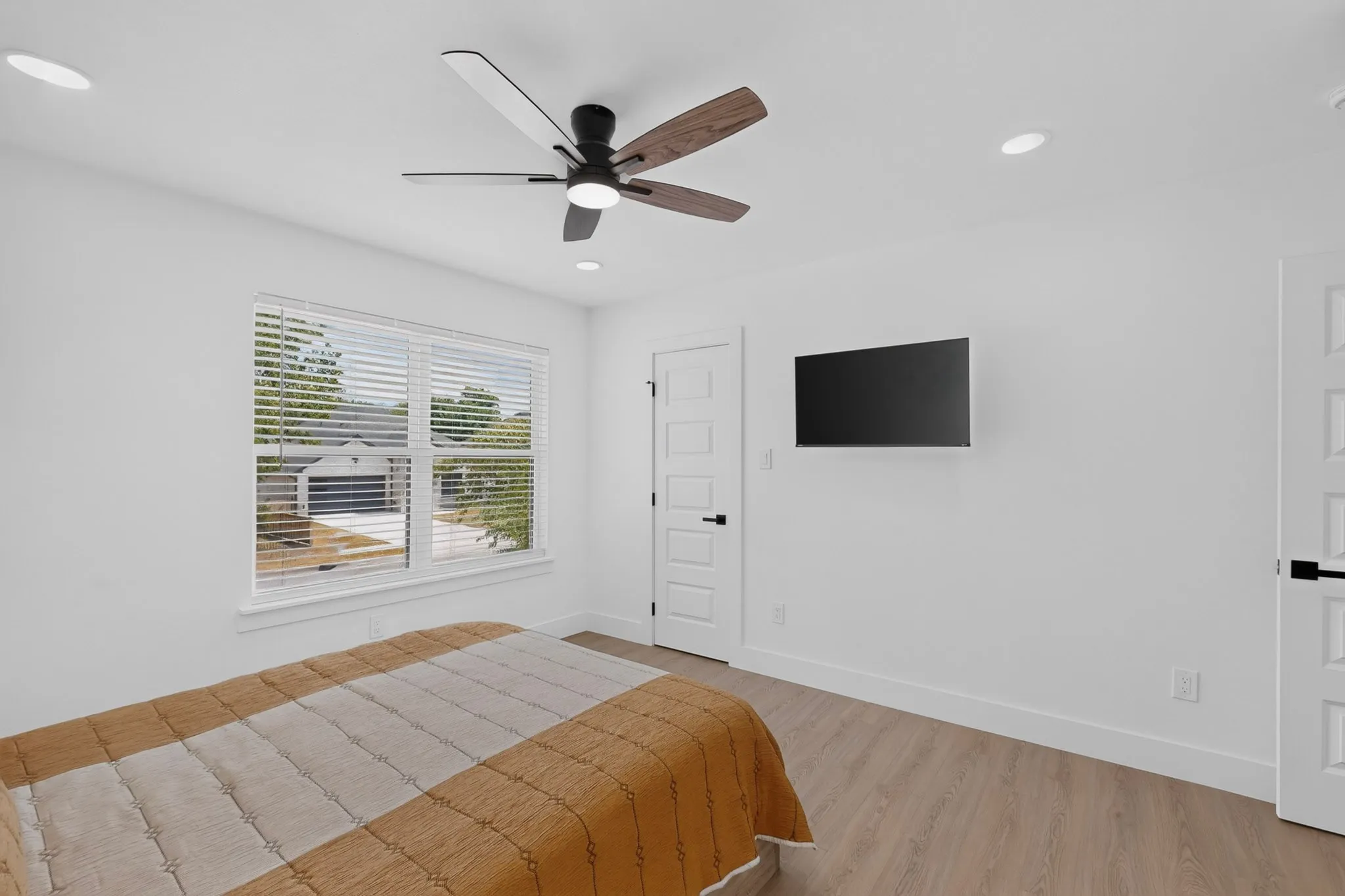 Wood-finish flooring, white paneled doors with black hardware, ceiling fan with integrated lighting, recessed ceiling lights, and window with blinds