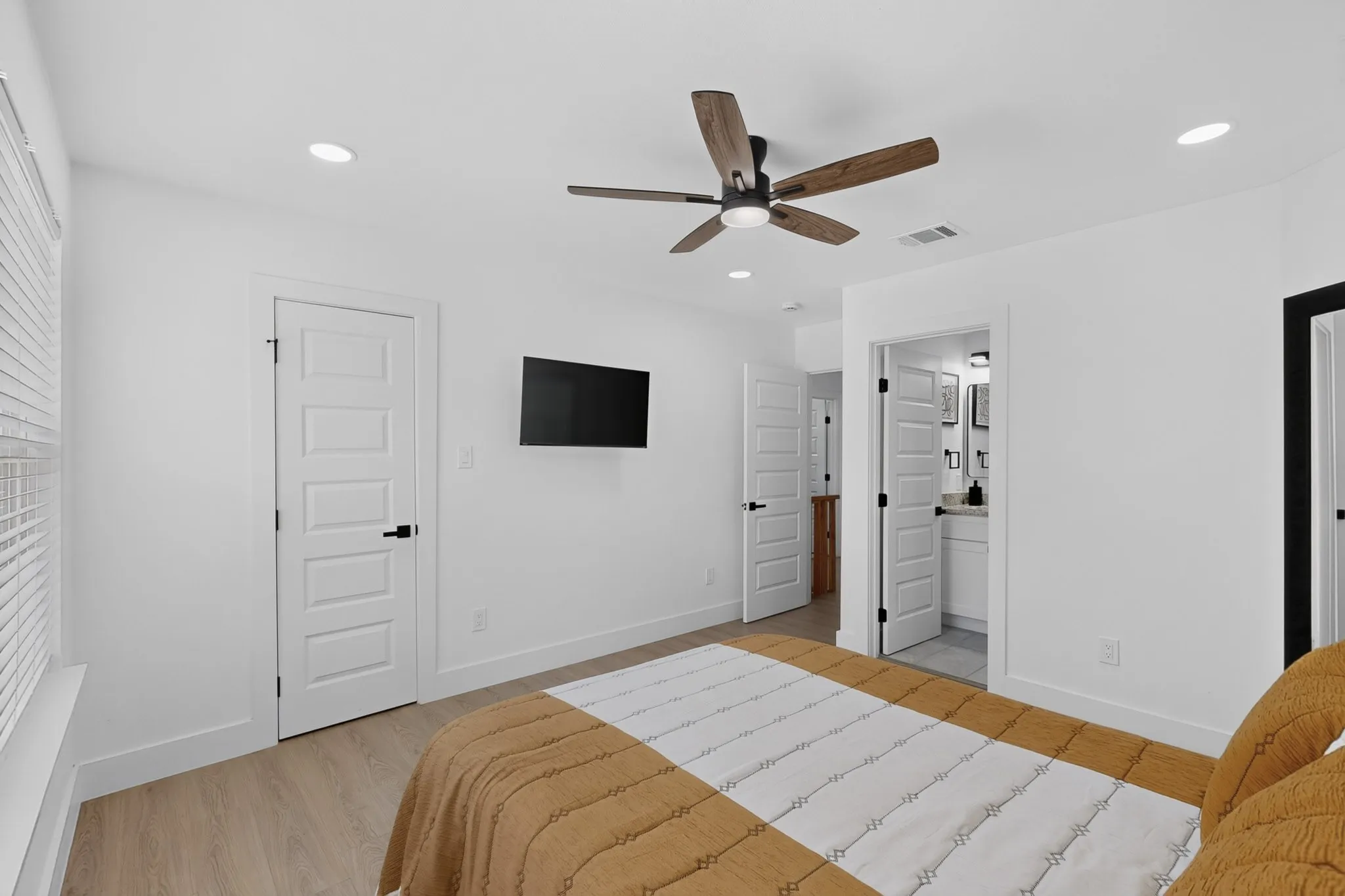 Recessed lighting, wood-finish flooring, ceiling fan with wood-finish blades, white paneled doors with black hardware, and an en-suite bathroom with a vanity and wall-mounted light fixtures
