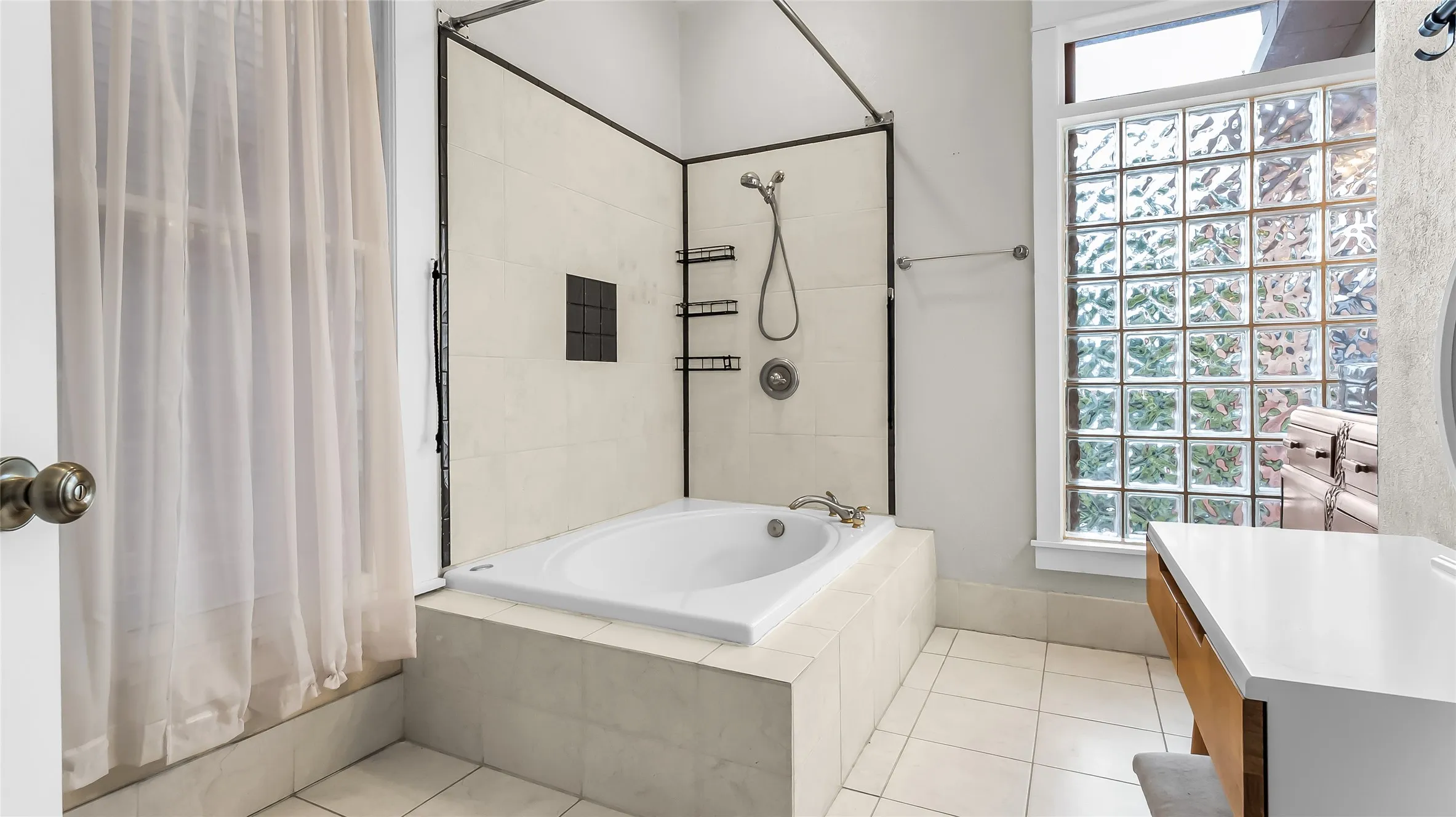 Soaking tub with a tiled surround, glass block window, and a white countertop vanity