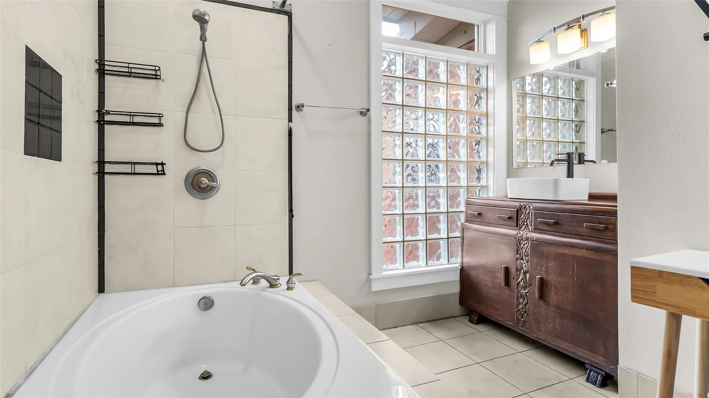 Built-in corner tub with tiled surround, glass block window, dark wood-finish vanity with an integrated vessel sink, and overhead lighting fixture